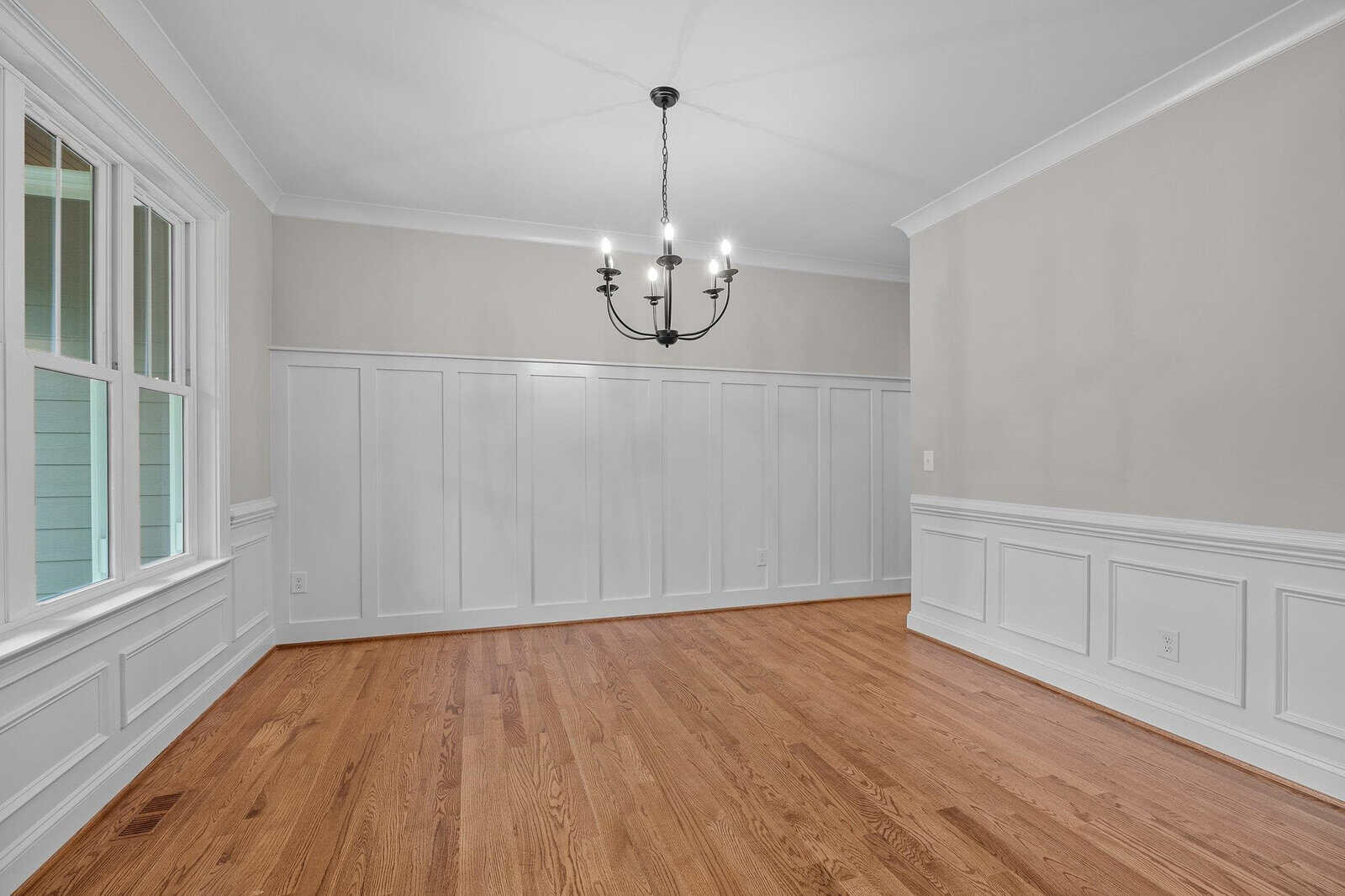 An empty room with white walls featuring wainscoting, a hardwood floor, and a black chandelier.