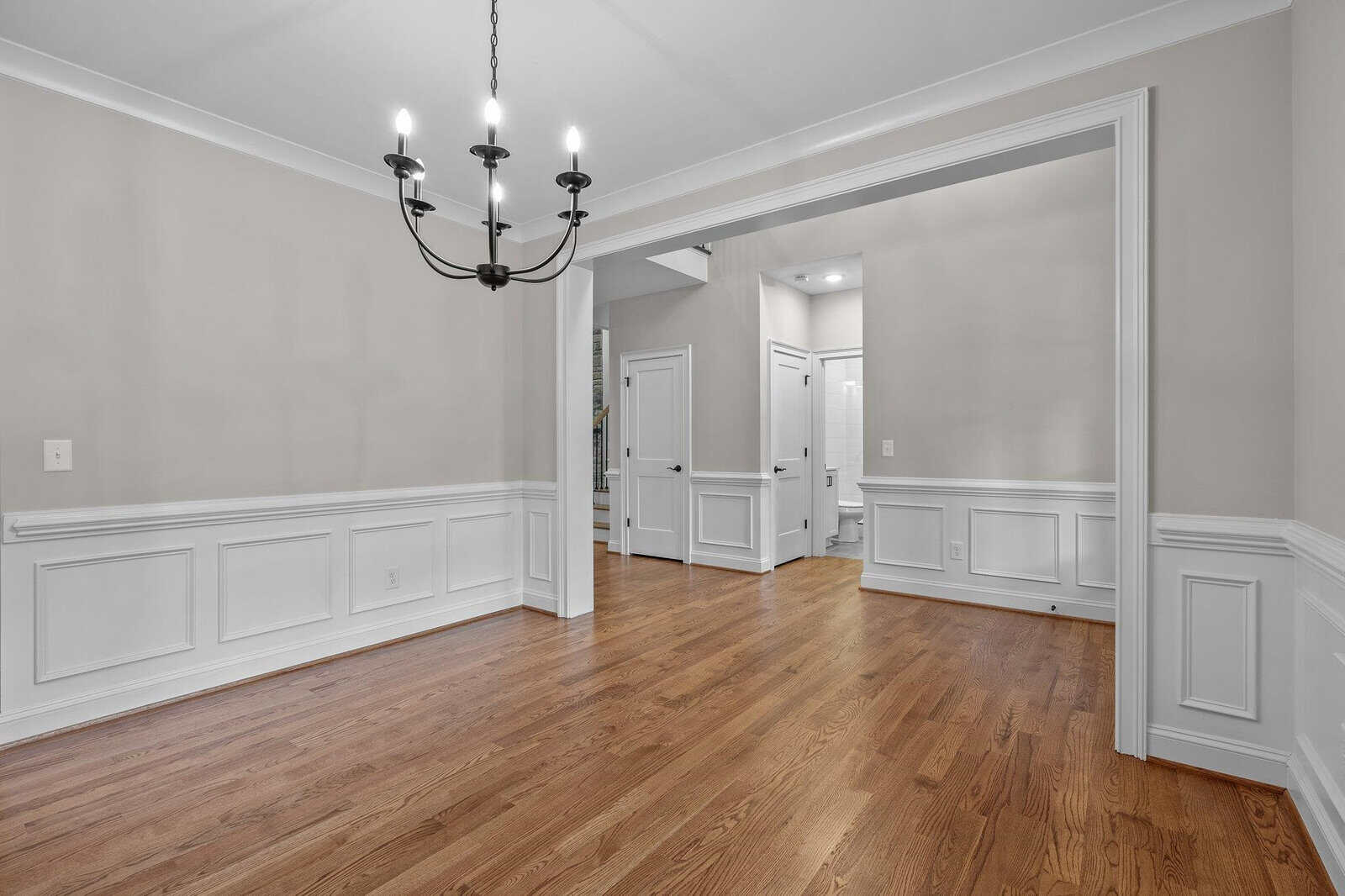 Empty room with hardwood flooring, white wainscoting, and a black chandelier, leading to another room through a wide doorway.