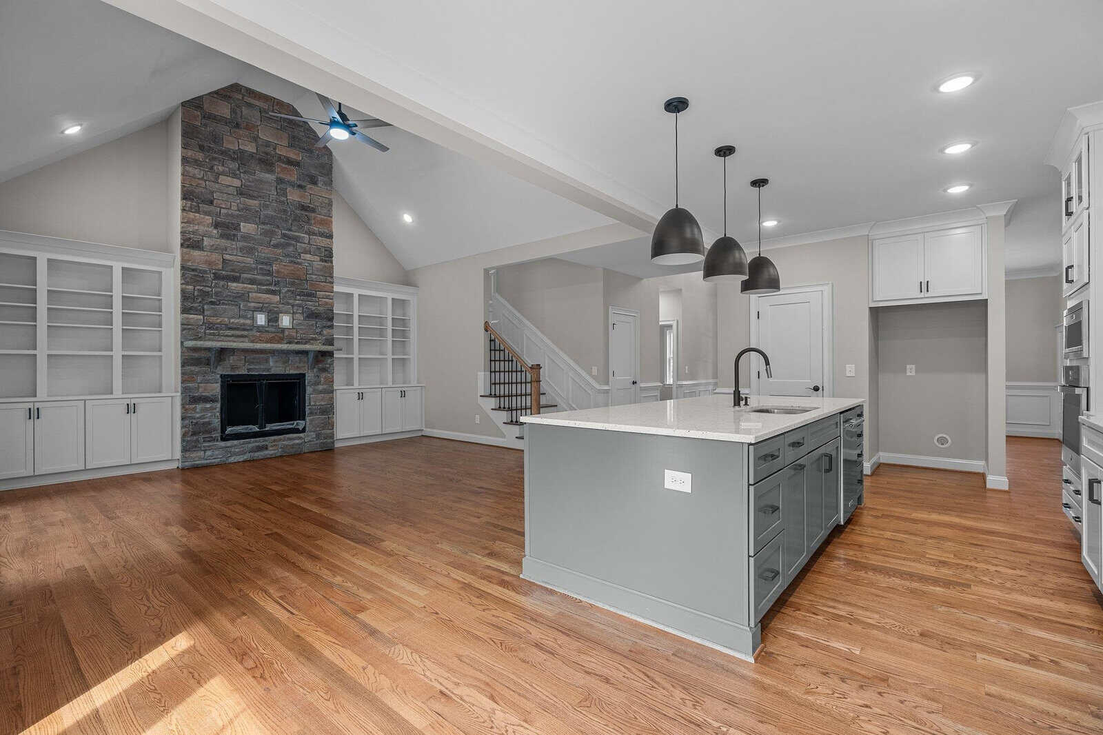 Modern spacious kitchen and living area with hardwood floors, white cabinetry, a central island with a sink, stainless steel appliances, a fireplace with a stone surround, and ample natural light.