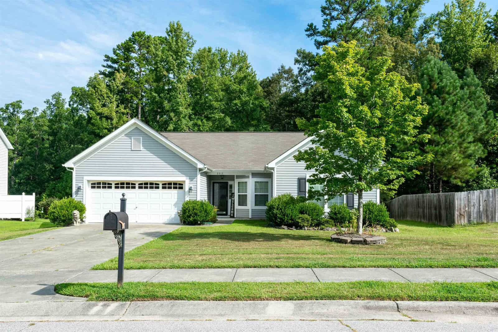 A charming single-story house at 2217 Bayswater Drive boasts light gray siding and a two-car garage. The manicured lawn, shrubs, and tree enhance its appeal. A black mailbox adorns the driveway, while Creedmoor's tall trees and clear blue sky provide a picturesque backdrop.