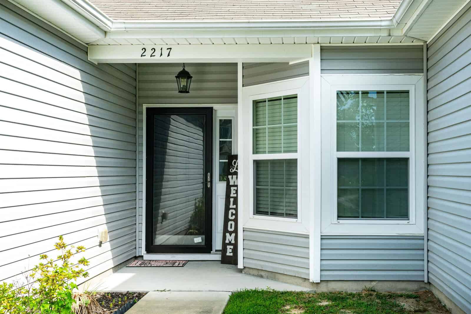 Front porch of a house with gray siding and a glass storm door. A welcome sign leans by the door, and the numbers 2217 are displayed above. Two windows with white frames are on the right, and a small garden is visible on the left.