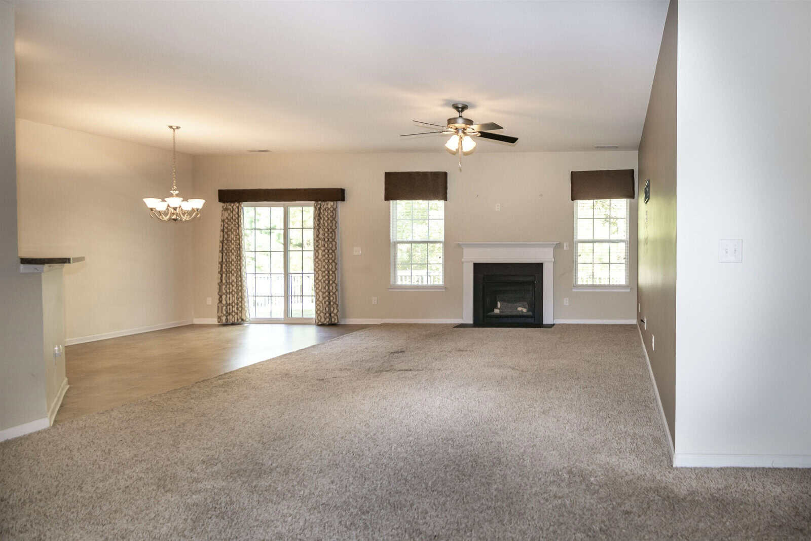 Spacious living room with beige walls and carpet. A ceiling fan is centered on the ceiling. The room connects to a dining area with a chandelier and has large windows with curtains, providing natural light. A dark fireplace is on one wall.