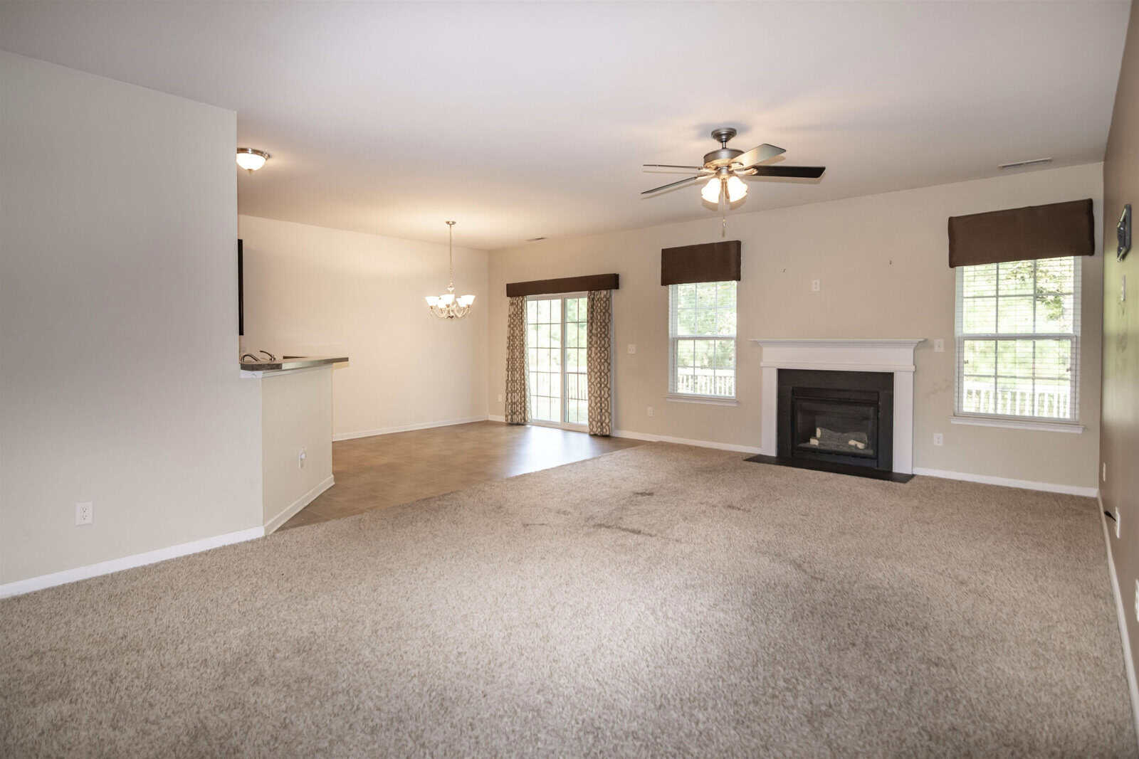A spacious, unfurnished living room with beige carpet and a tiled dining area. It features a ceiling fan, fireplace, and large windows with valances, leading to a patio door with curtains. The walls are painted a light cream color.