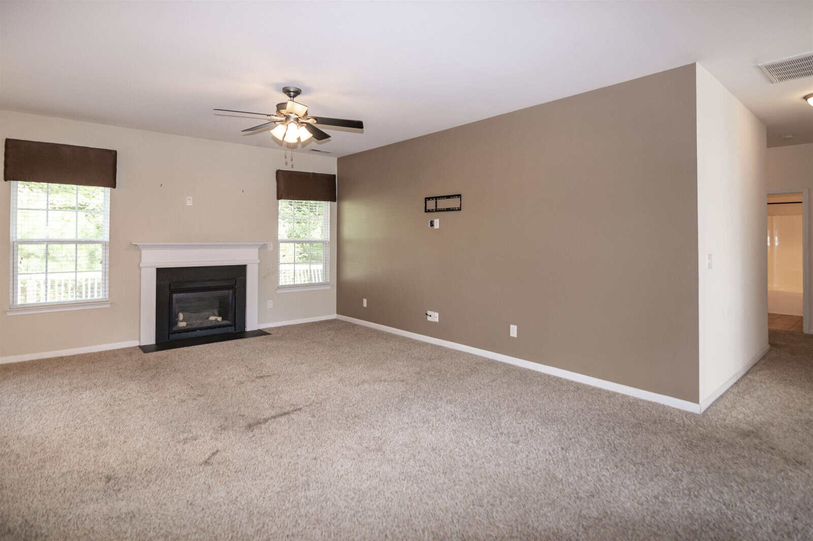 A spacious living room with beige carpet and neutral walls featuring a ceiling fan and a fireplace. Two windows with valances are on the left wall. A white door is partially open to the right. The room is unfurnished.
