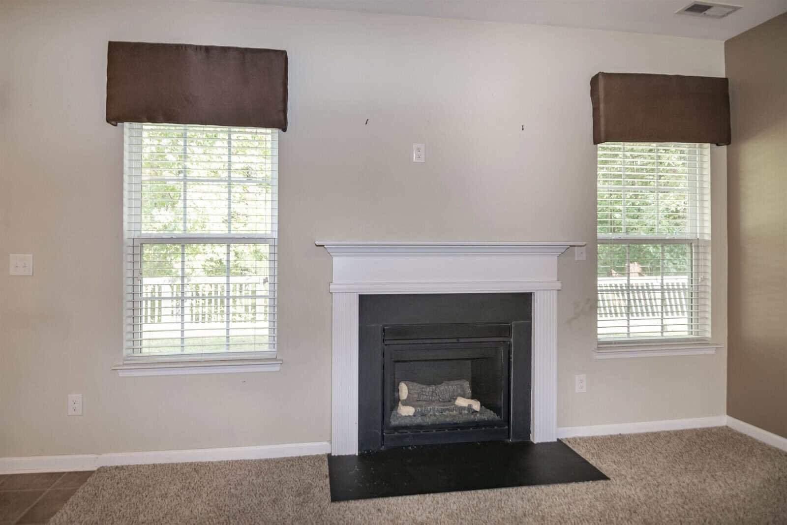 A living room with a beige carpet and walls, featuring a fireplace with a simple white mantel. Two large windows with brown valances flank the fireplace, letting in natural light and offering a view of a backyard fence.