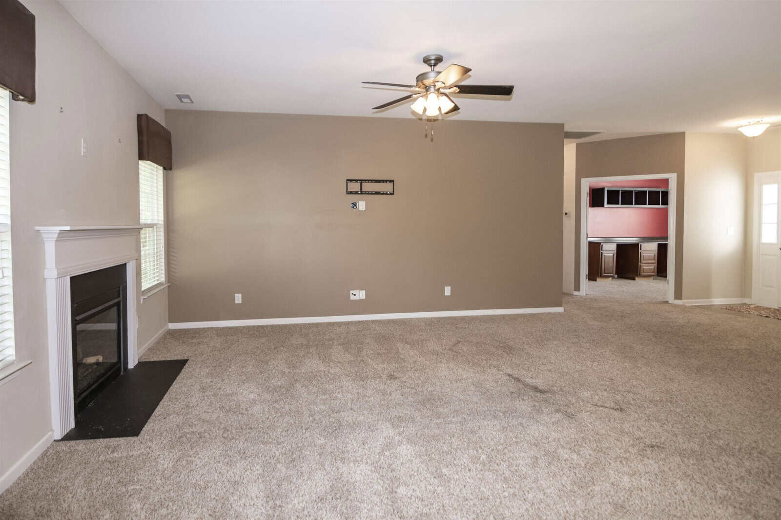 Spacious living room with neutral walls and carpet, featuring a ceiling fan with lights. There's a white fireplace on the left and two windows. An open doorway leads to another room with distinctive red walls and dark wood trim.