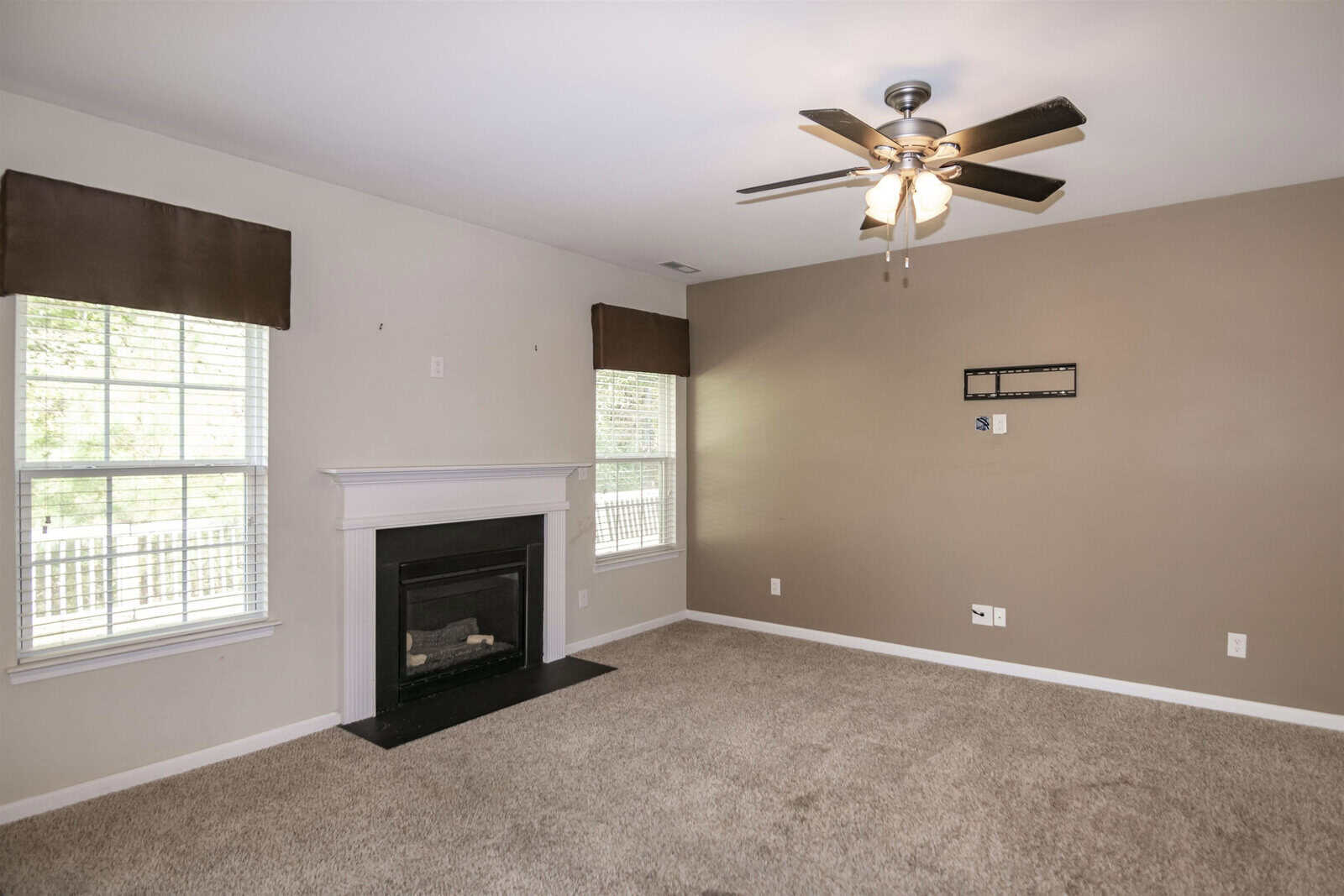 A living room with light beige walls and carpeted floor. It has a ceiling fan with lights, a white mantel fireplace on the left wall, and two windows with brown valances. A wall mount for a TV is centered on the right wall.