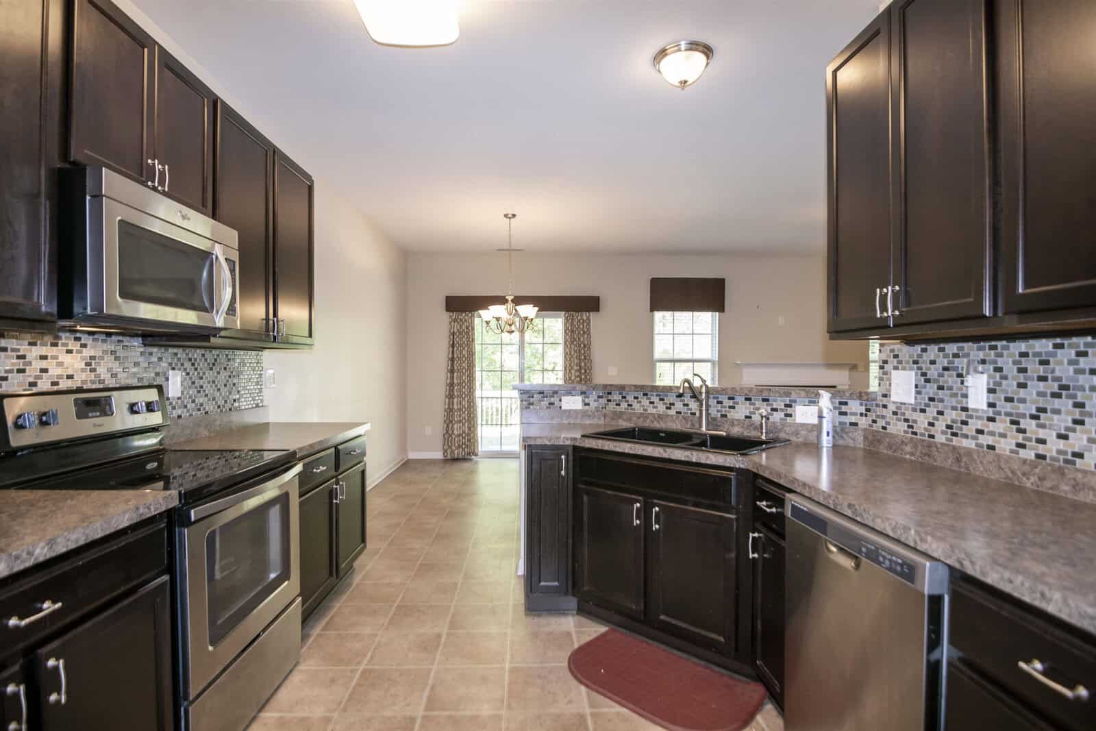 A modern kitchen featuring dark wooden cabinets, stainless steel appliances, and a tile backsplash. The room is highlighted by overhead lighting, a central island with a sink, and a view into a dining area with large windows in the background.