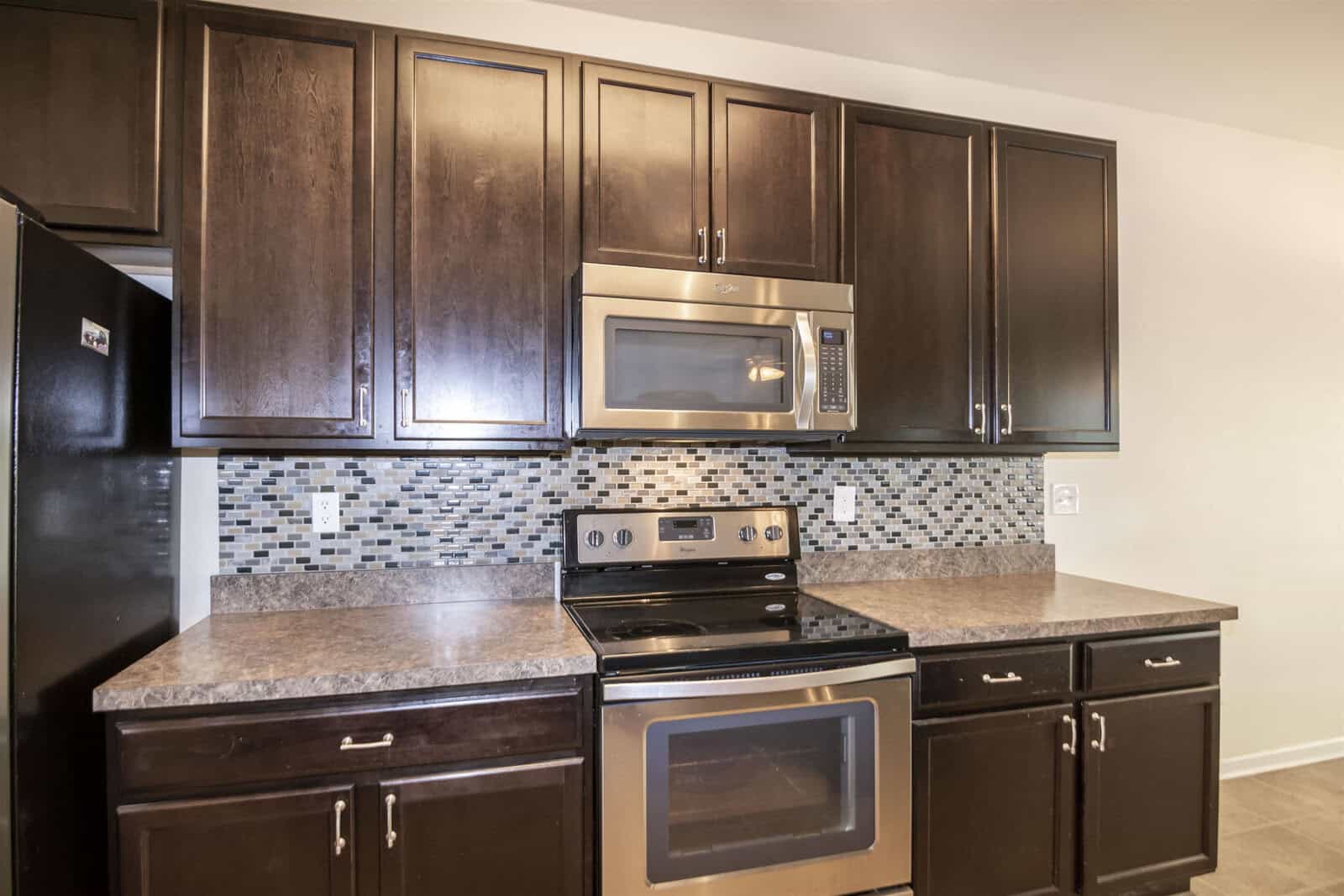 A modern kitchen featuring dark wood cabinets, a stainless steel oven with a stovetop and microwave, and a gray speckled countertop. The backsplash is a mosaic of gray, black, and white tiles.