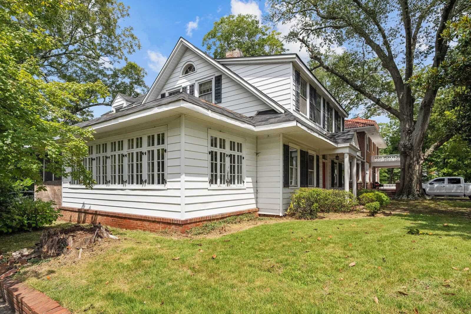 A two-story white clapboard house with black shutters, featuring a covered front porch, surrounded by a lawn and mature trees under a partly cloudy sky.