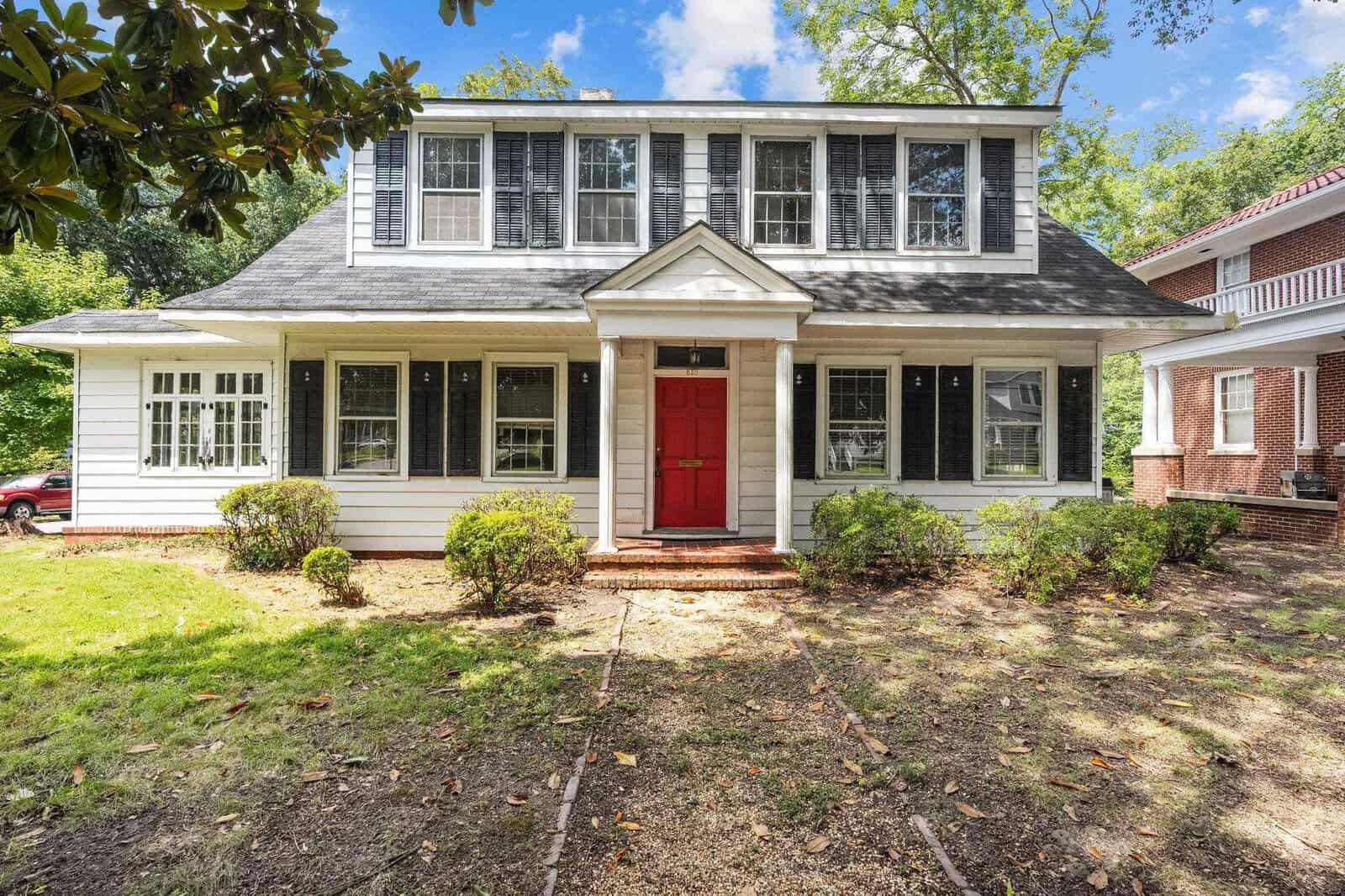 Two-story house with white siding and a red door, flanked by large windows with shutters, and surrounded by greenery under a partly cloudy sky.