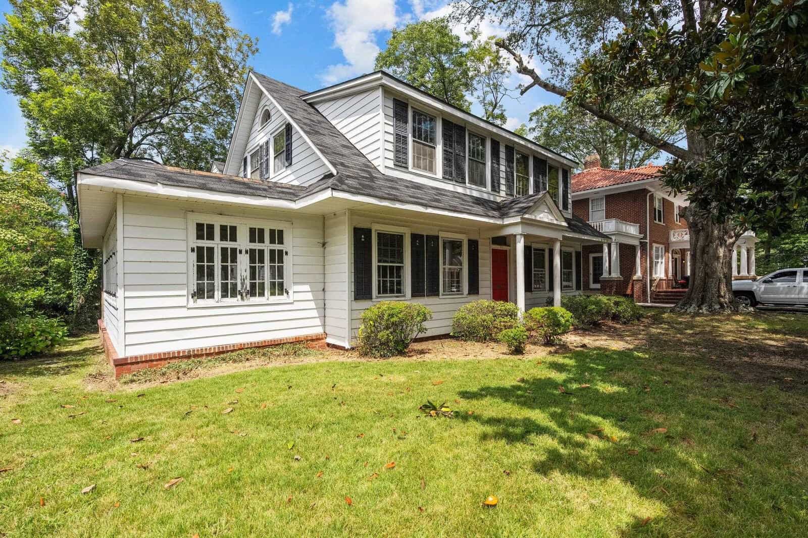 A two-story suburban house with white sidings and black shutters, featuring a red door and surrounded by a well-maintained lawn with scattered trees.