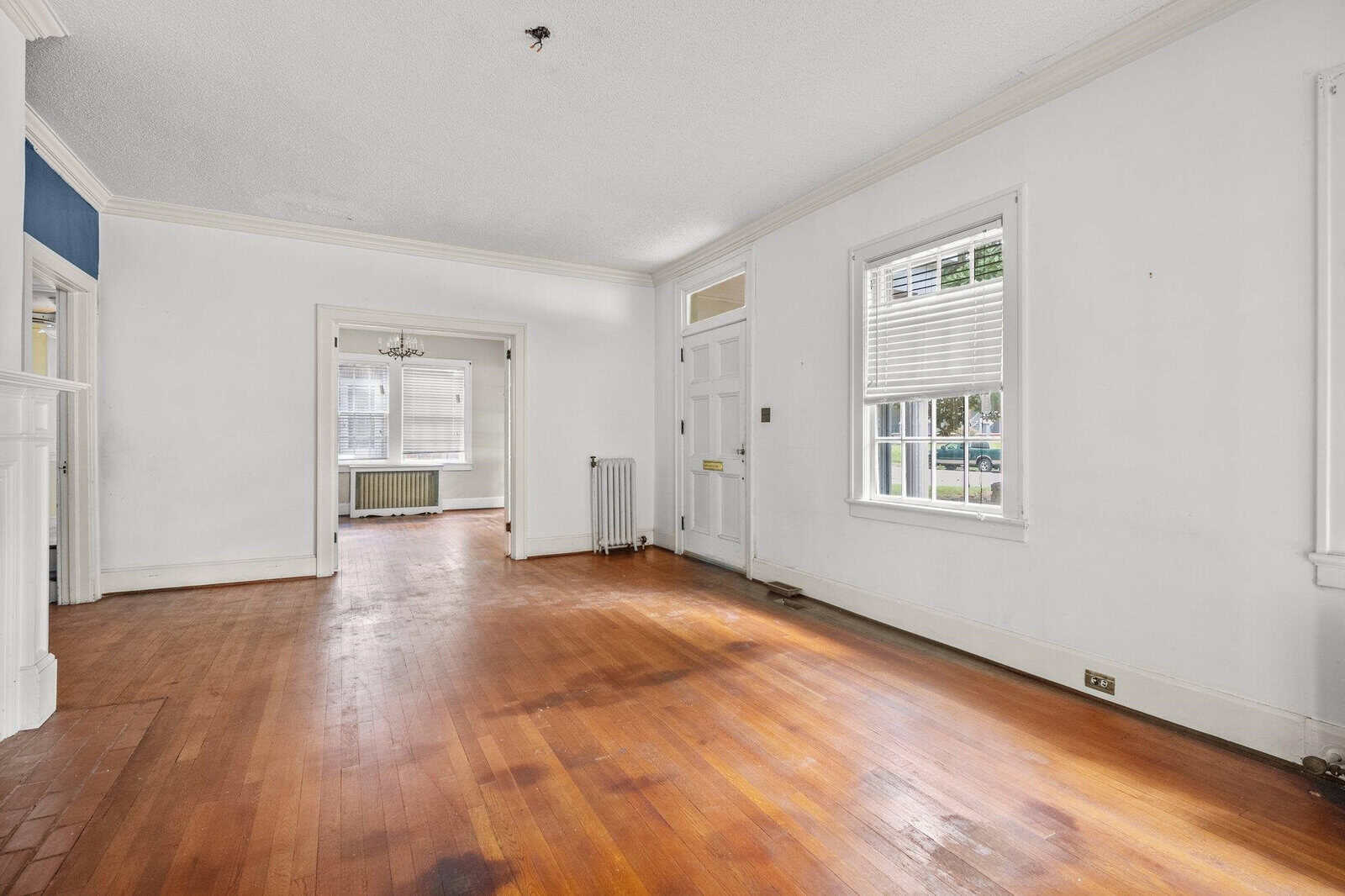 Empty living room with hardwood floors, white walls, and a white door at the far end, featuring one window to the right and a staircase to the left.
