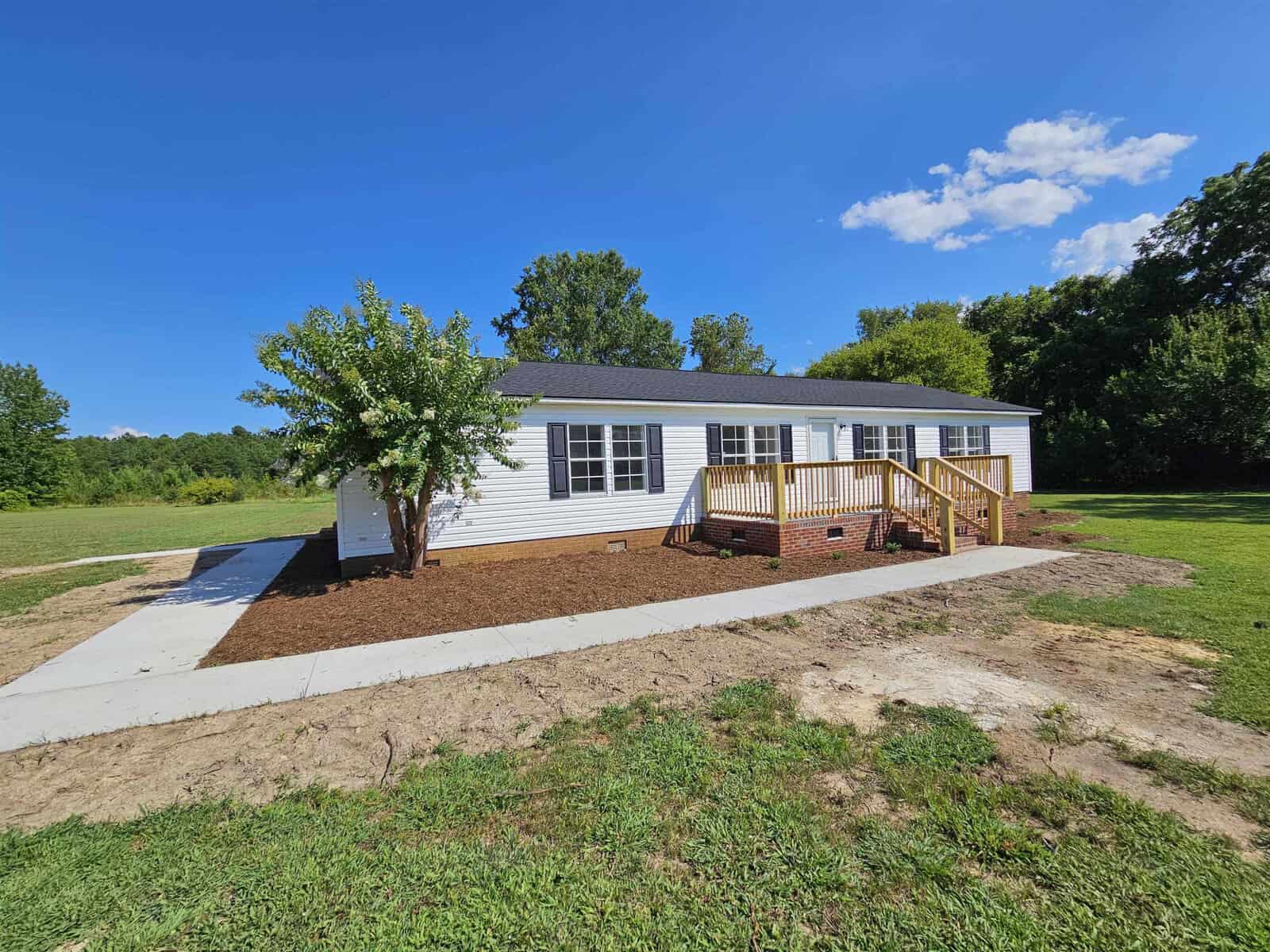 A small white house with a gray roof and a wooden porch sits on a grassy lot under a clear blue sky. A tree is on the left side of the house, and a concrete path leads to the porch. Surrounding trees are visible in the background.