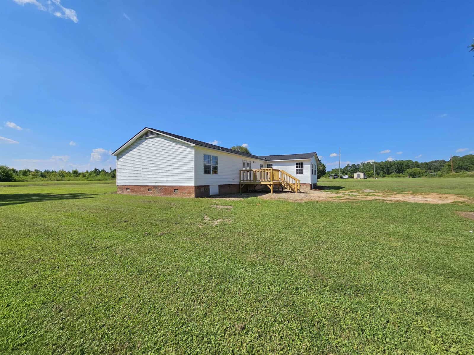 Wide view of a single-story white house with a dark roof, wooden ramp, and brick foundation, set on a large grassy field under a clear blue sky.