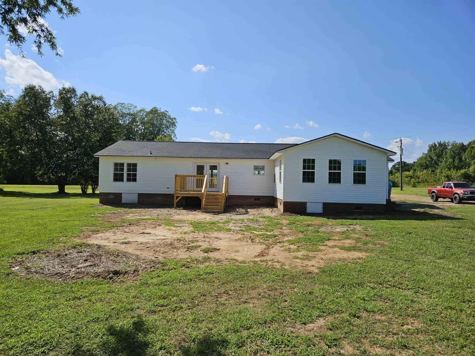 A white single-story house with a small wooden deck and stairs leading to a grassy yard. A red truck is parked on the right side, and trees are visible in the background under a clear blue sky.