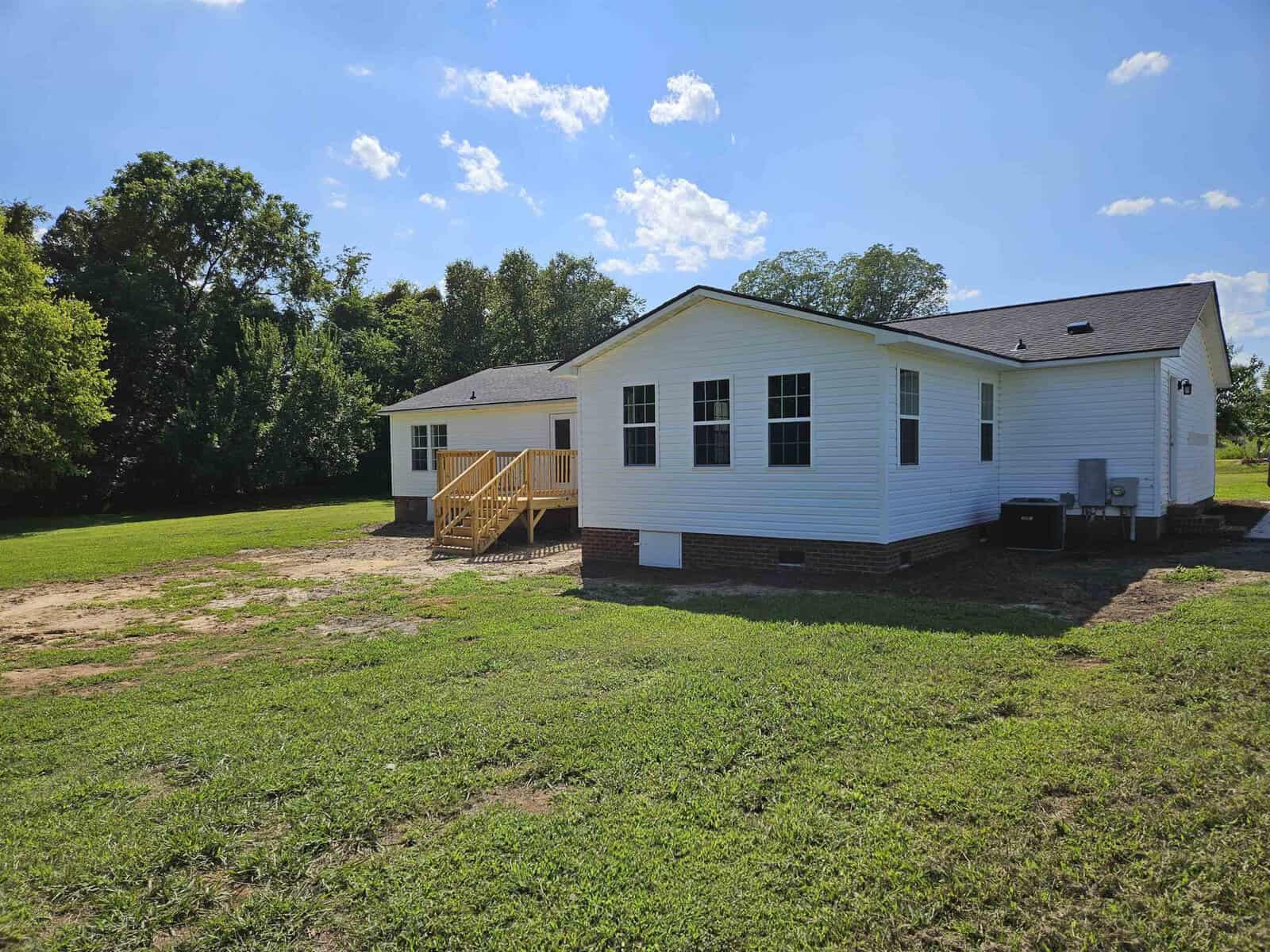 A white, single-story house with a gray roof and a wooden deck at the back, set on a grassy lawn with trees in the background under a clear blue sky.