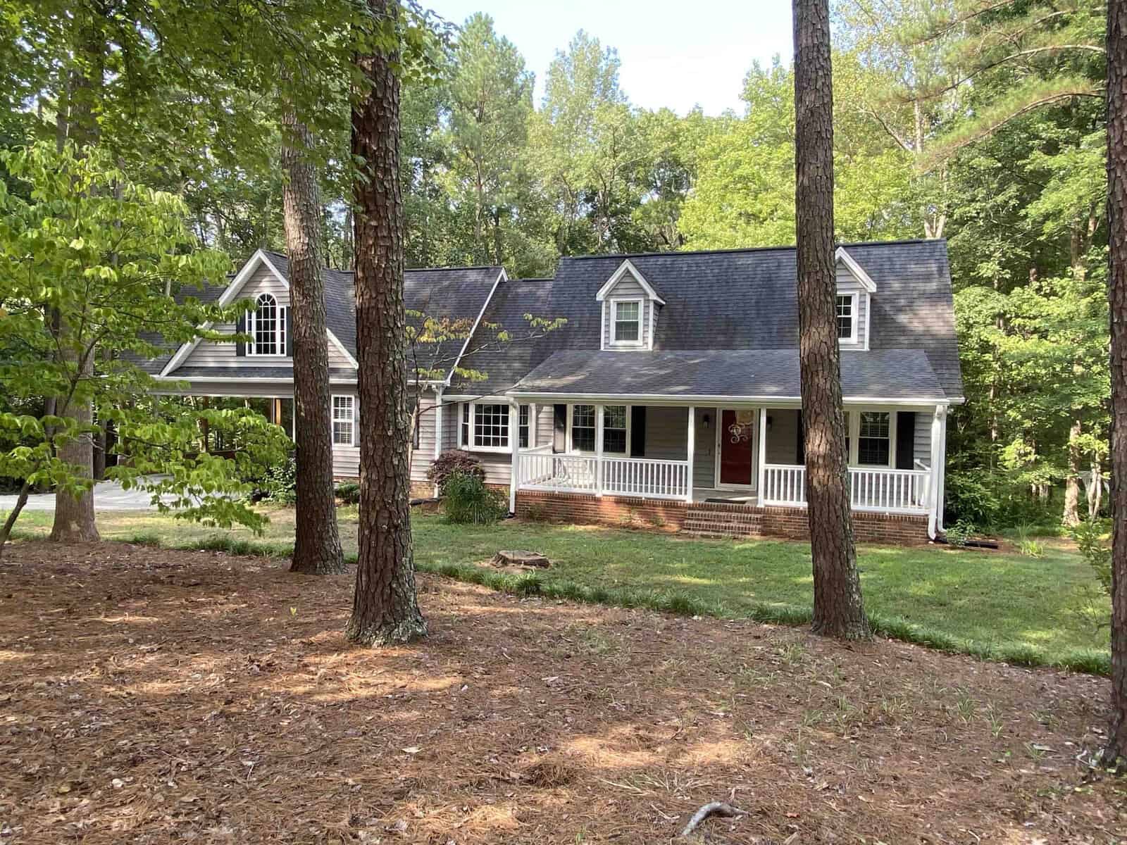 A single-story house with a gabled roof and a front porch, surrounded by trees with a partly shaded lawn.