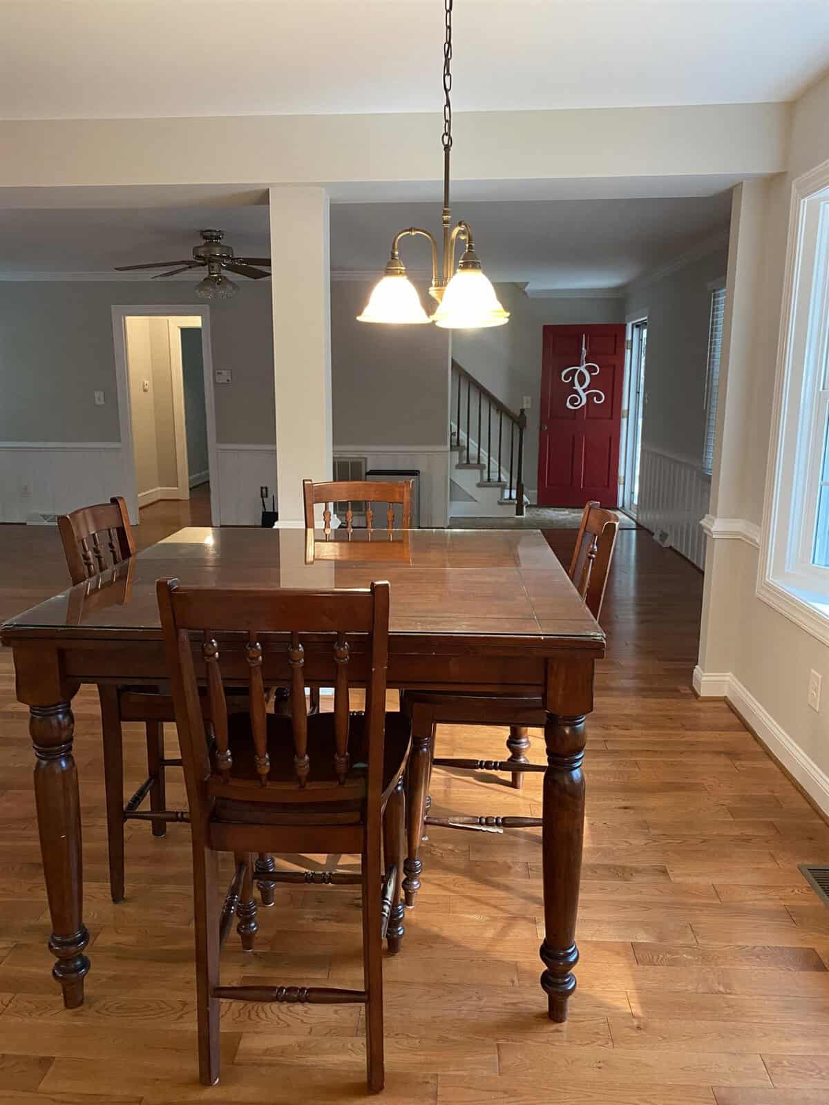 A wooden dining table with chairs set in a well-lit room with hardwood floors, white walls, and a red door in the background.