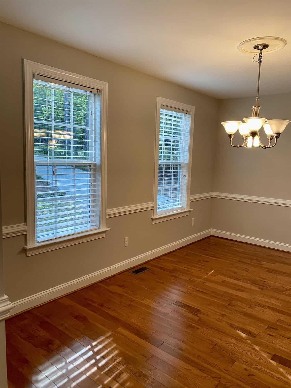 An empty room featuring polished hardwood floors, two windows with white blinds, and a ceiling light fixture, with a view of the trees outside.