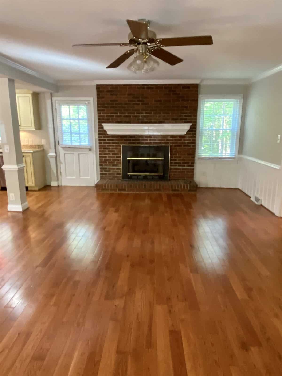 Empty living room with a brick fireplace, hardwood floors, white walls, and a ceiling fan.