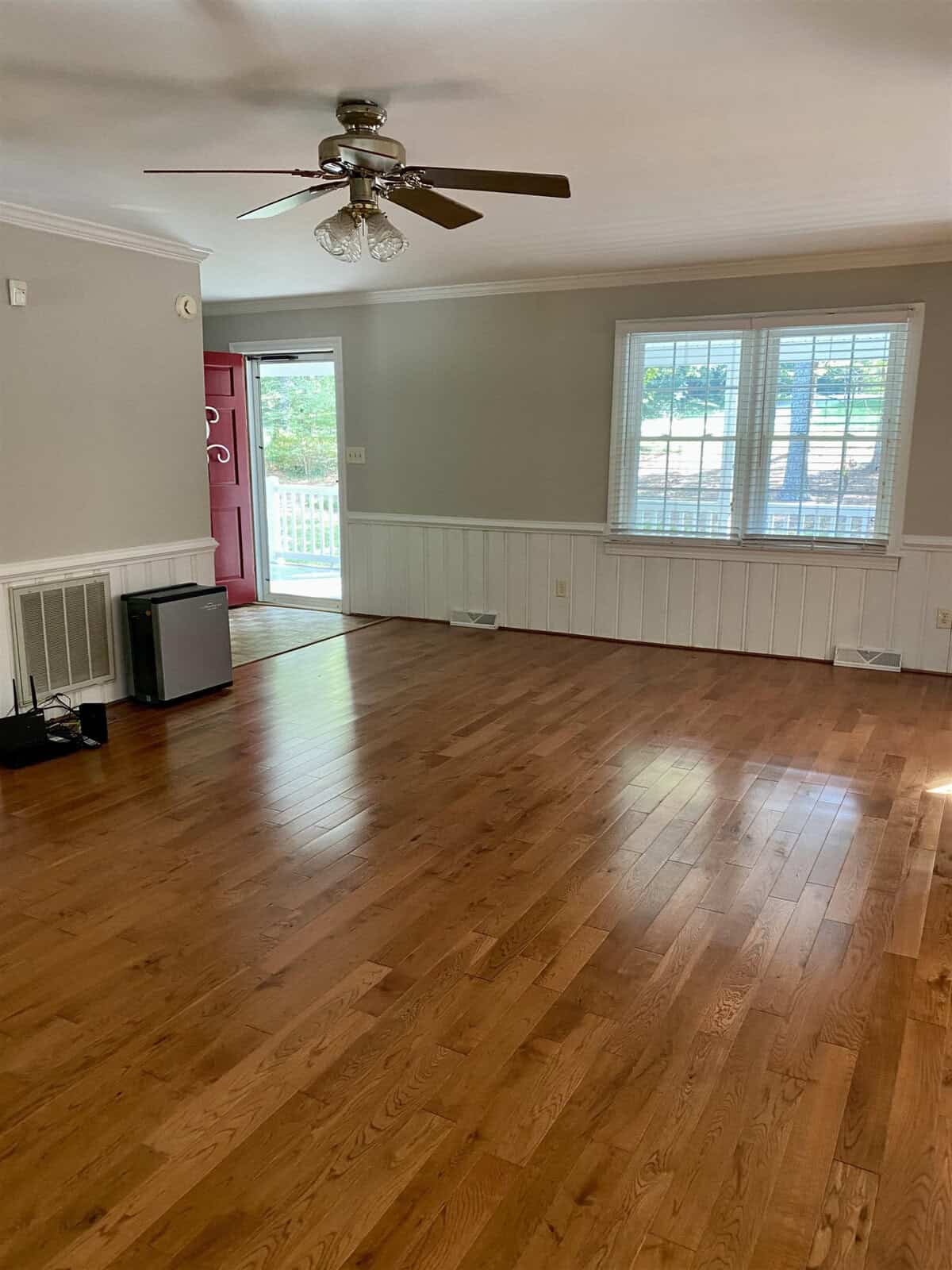 An empty room with hardwood floors, white walls with wainscoting, a ceiling fan, and large windows with a view of the outside, leading to a deck with a pink door to the left.