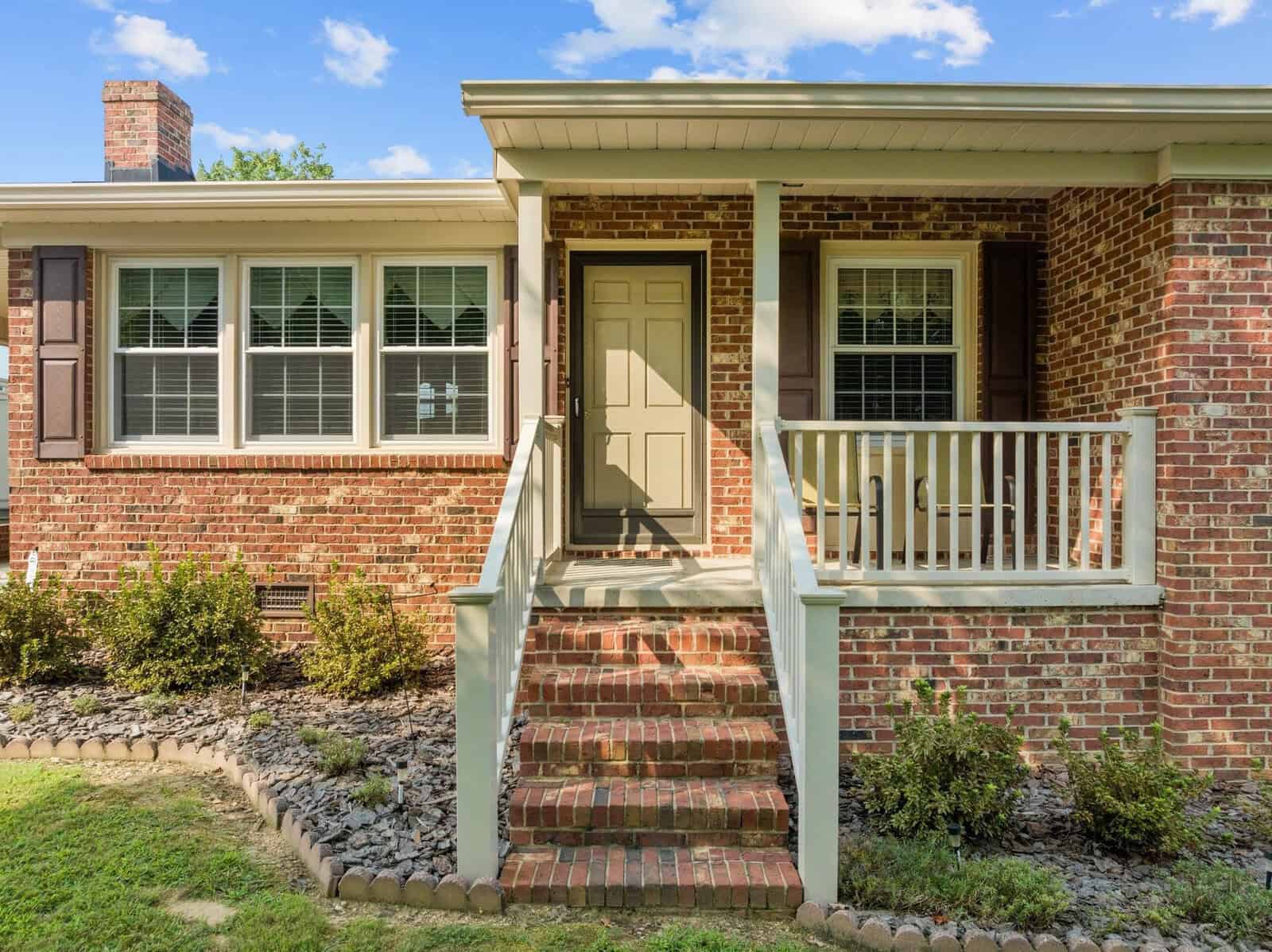 A brick house with a beige front door, white railing, and steps leading up. Windows with shutters are on either side of the door. Small shrubs and a stone border frame the house, and the sky is partly cloudy.