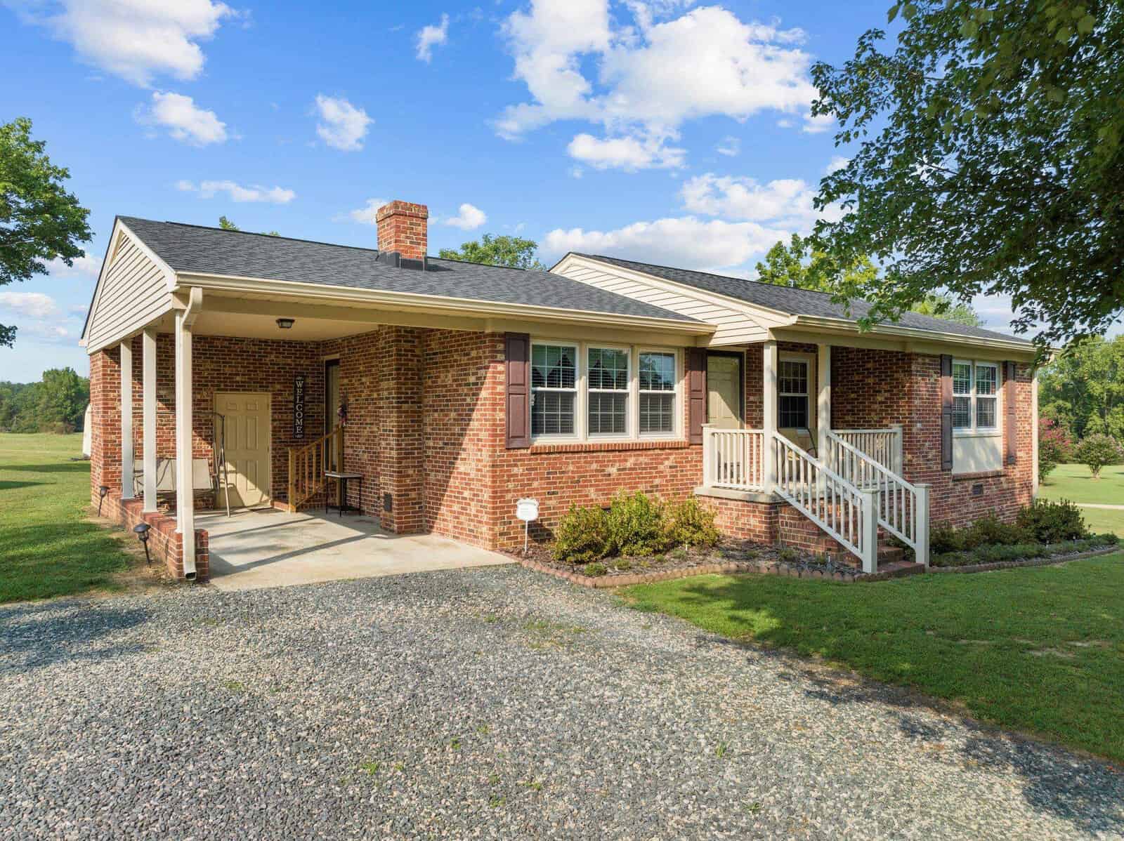 A single-story brick house with a front porch, white railing, and a carport. It is surrounded by a grassy yard and trees under a blue sky with scattered clouds. Gravel driveway leads to the carport.