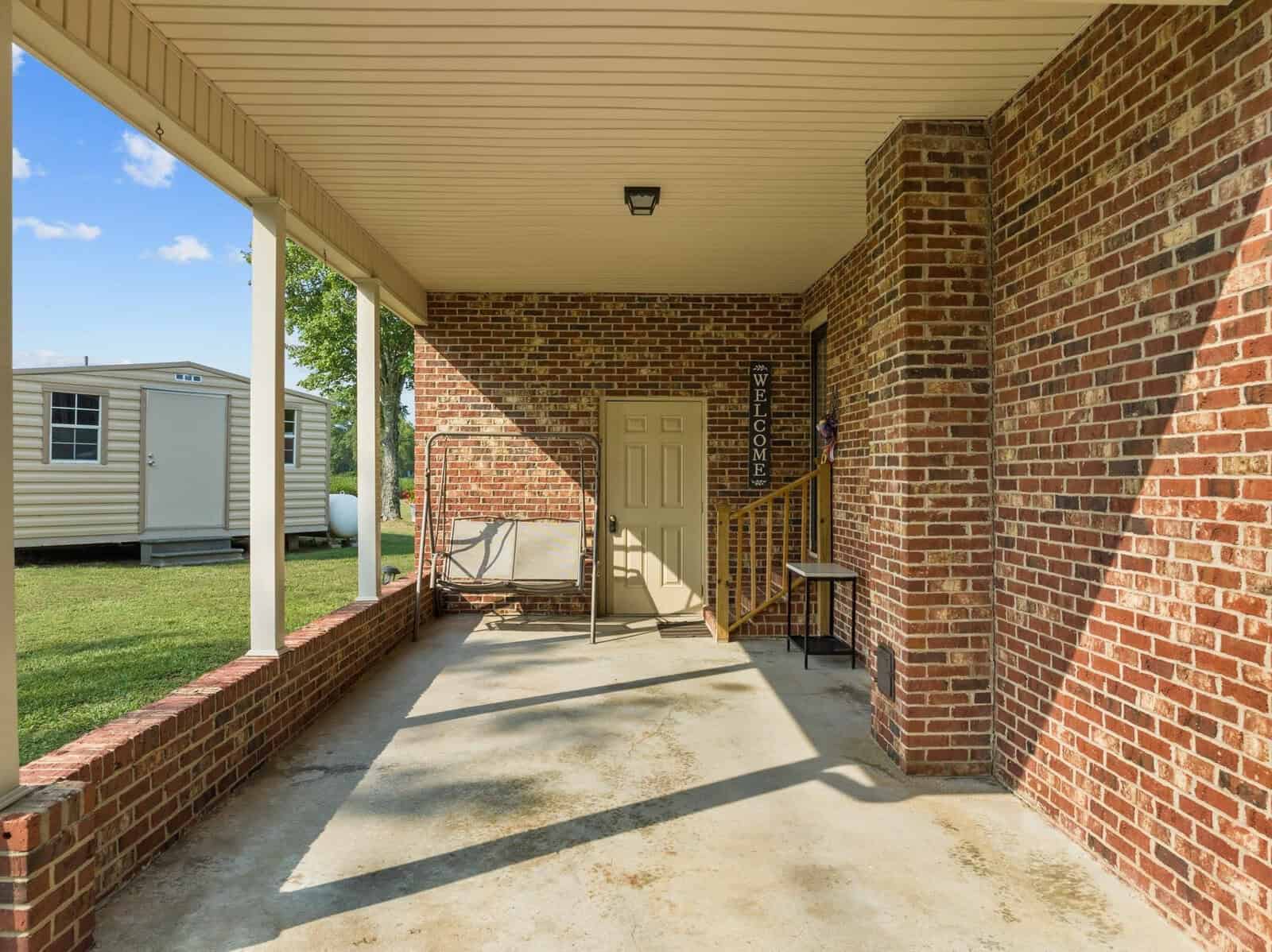 Covered patio area with brick walls and concrete floor. A swing chair is on the left, and a welcome sign is next to a door on the right. Steps with a black railing lead up to the door. A small yard and storage shed are visible outside.