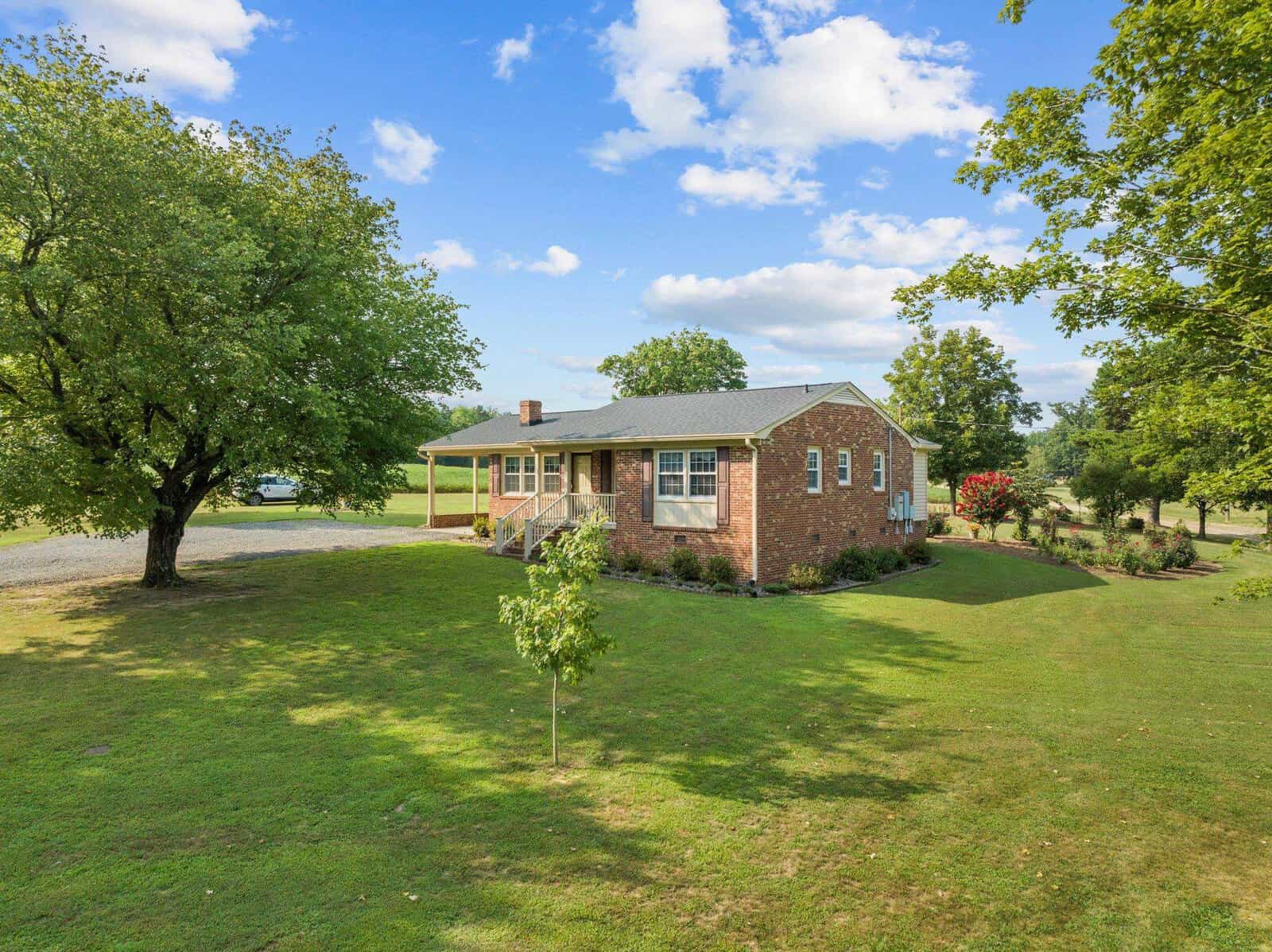 A single-story red brick house with a gray roof, surrounded by a lush green lawn and trees. The sky is clear with scattered clouds, and a gravel driveway leads to the house. A few shrubs and a small tree are in the front yard.