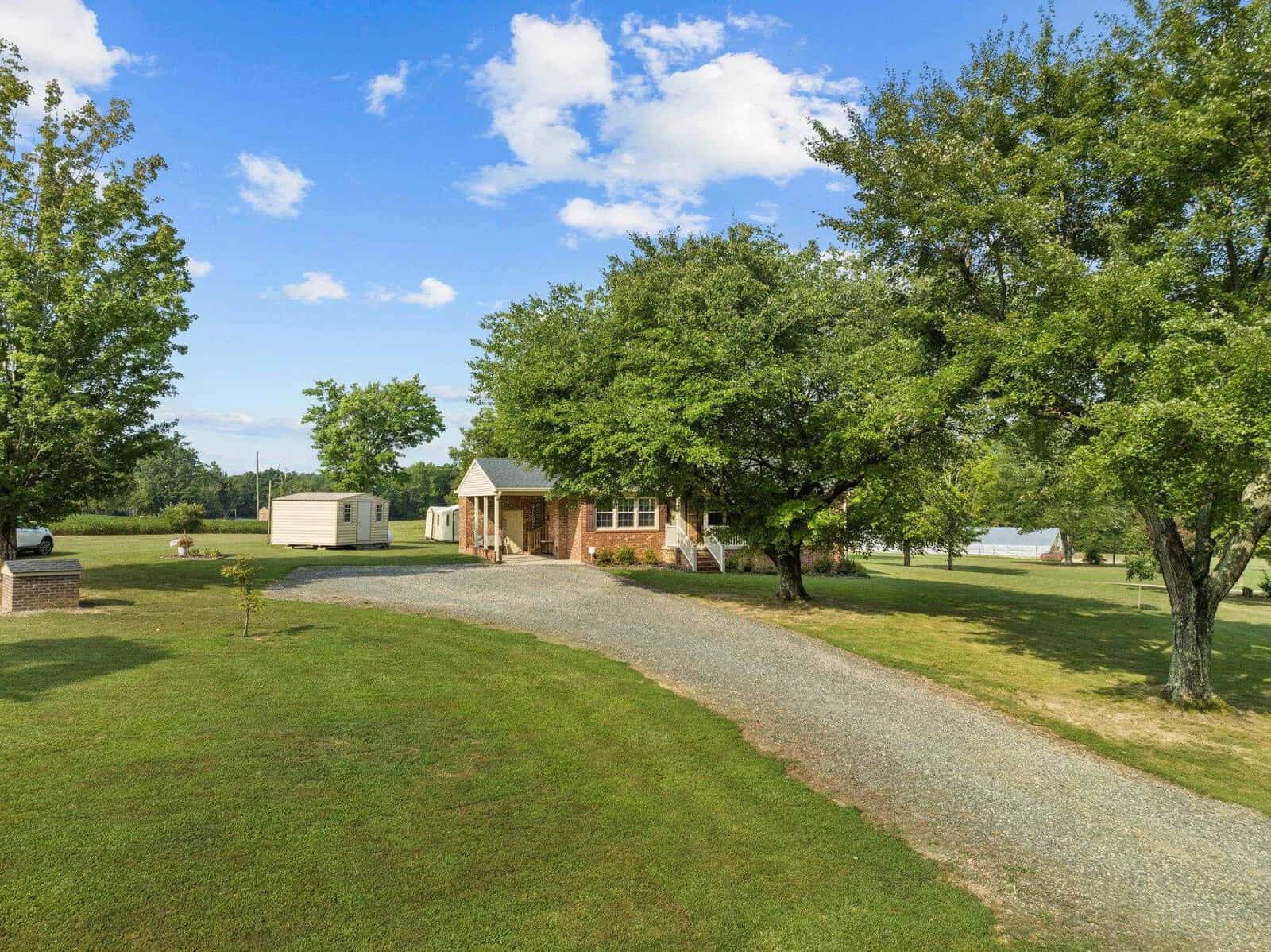 A red brick house with a gray roof sits in a lush, grassy yard. A gravel driveway curves towards the front entrance. Mature trees provide shade, and a small shed is visible in the background under a partly cloudy sky.