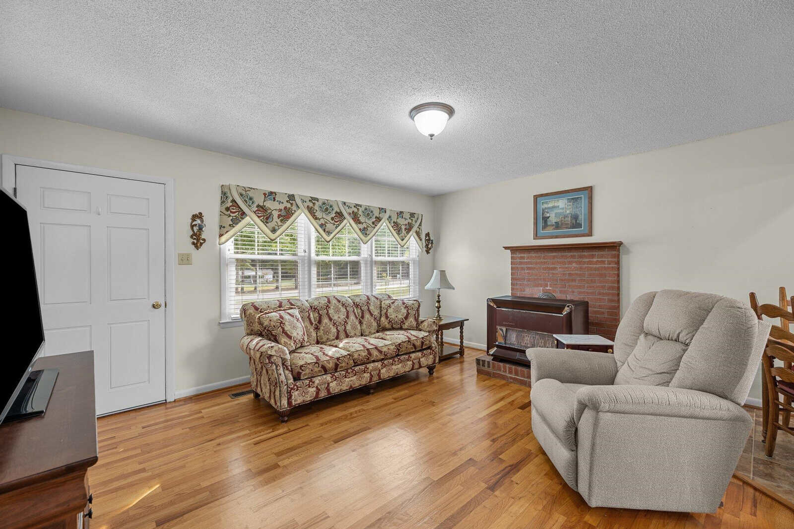 A cozy living room features hardwood floors, a floral-patterned sofa, a beige recliner, and a TV. A painting hangs above a small brick fireplace. A wooden dining table with chairs is visible. Large windows are adorned with patterned valances, allowing natural light.