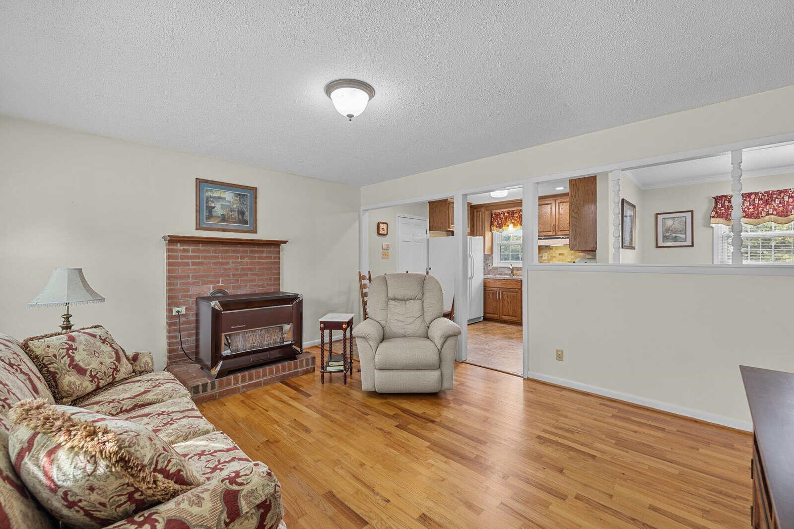 A cozy living room featuring a floral-patterned sofa, an armchair, and a brick fireplace with a framed artwork above. The room has wooden flooring and opens into a kitchen area, visible in the background through a cut-out wall.