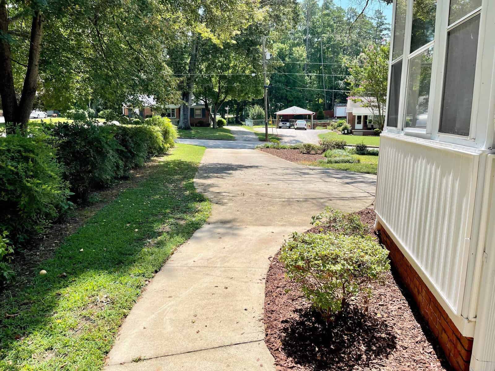 A sunny day view of a residential area with a concrete pathway leading away from the foreground, bordered by green shrubs and a white structure to the right. trees and homes are visible in the background.