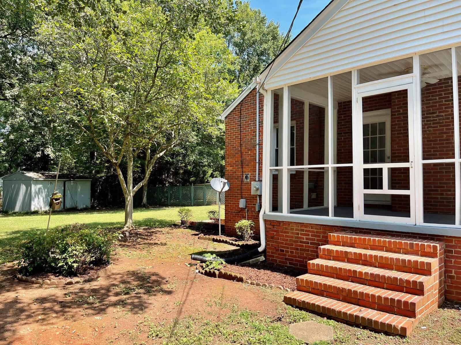 A sunny day shining on the side of a brick house with a screened porch and a well-kept lawn featuring a tree, some shrubs, and a satellite dish.