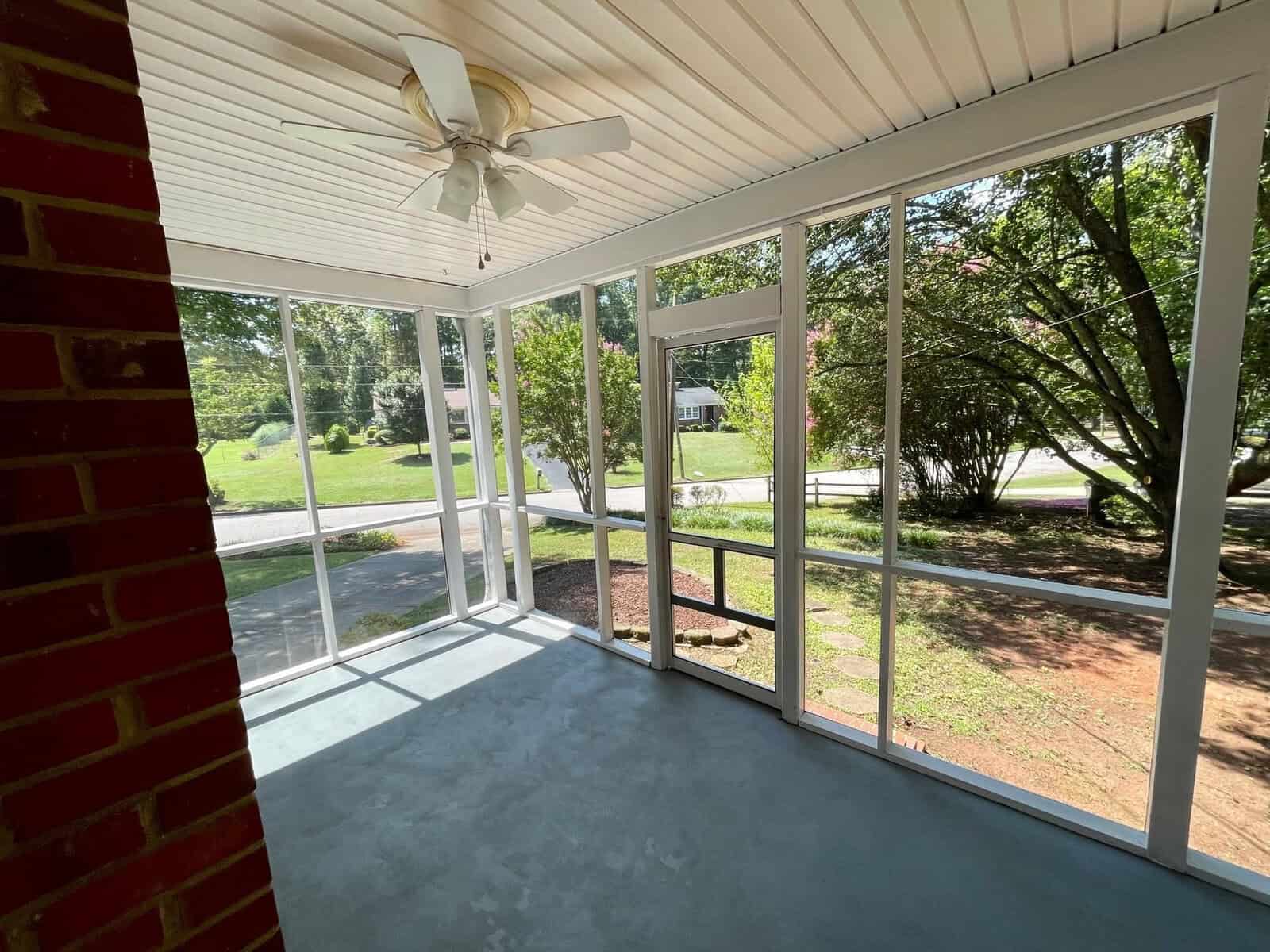 A sunlit screened porch with a ceiling fan, overlooking a residential street with greenery.