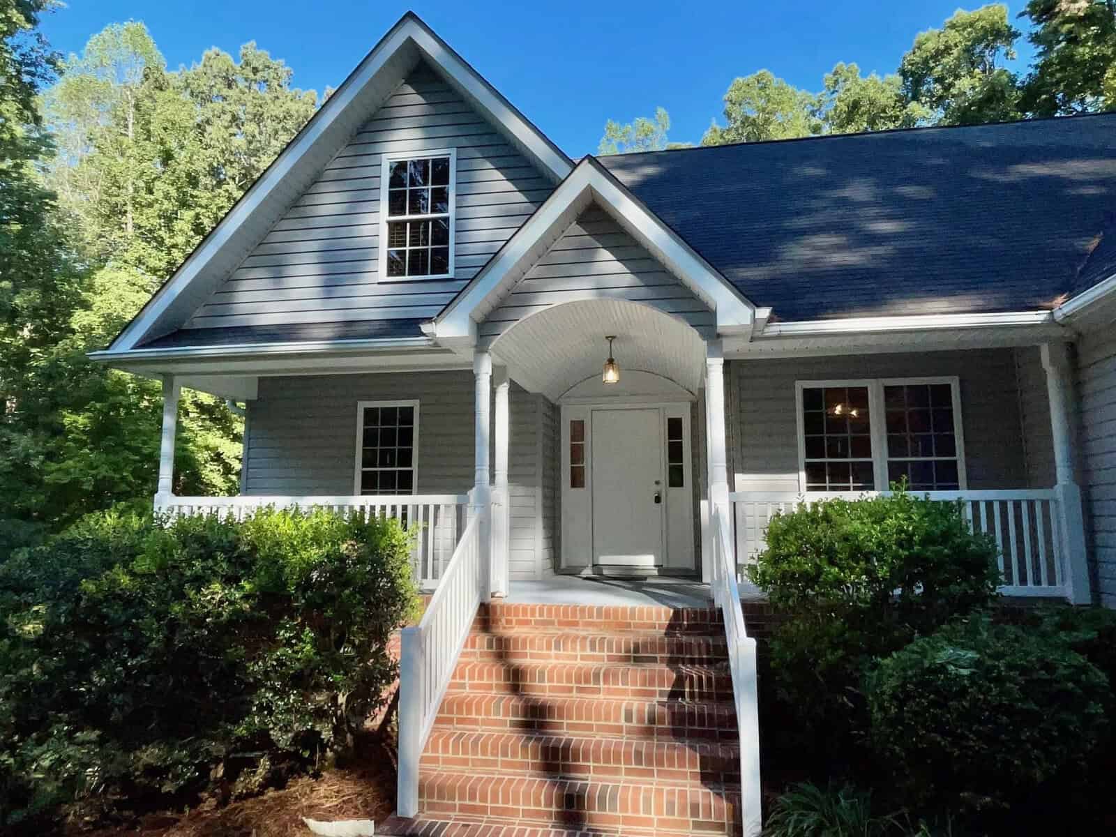 A quaint gray house with white trim, featuring a brick staircase leading up to a covered front porch nestled amongst green trees.