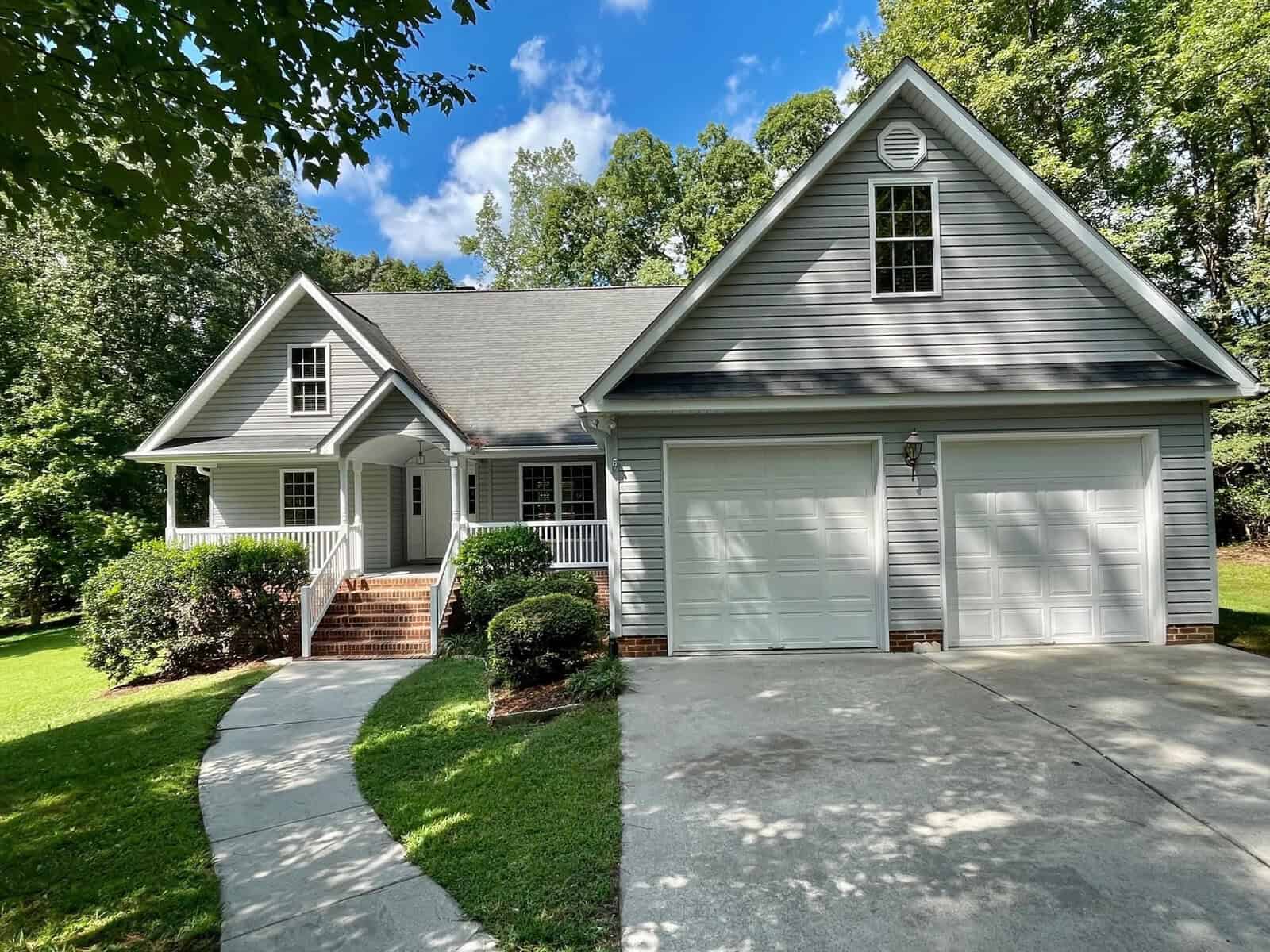 A single-story house with a gabled roof, a double garage, and a front porch surrounded by trees and greenery under a clear sky.