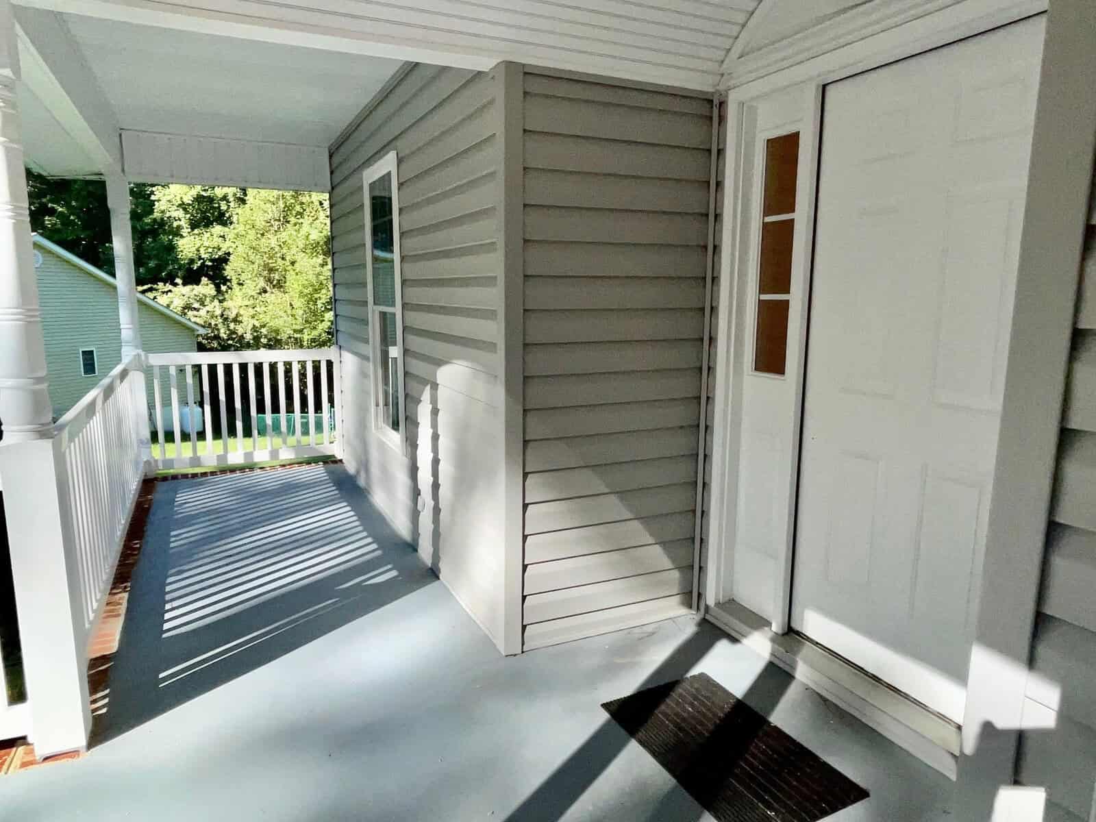 A sunlit porch with shadows cast on the floor, featuring a white front door partially open to the left, a doormat, and railing along the exterior.