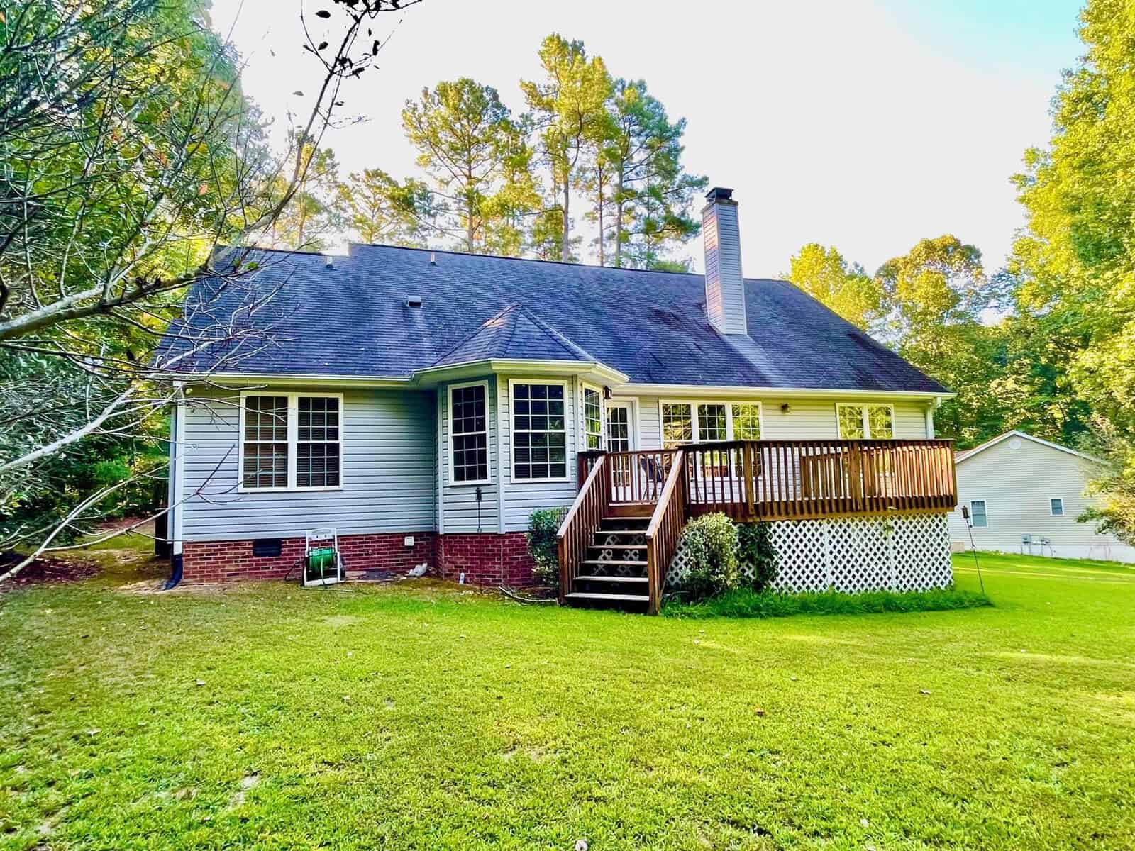 Single-story house with gray siding and a wooden deck, surrounded by green lawn and trees under a clear sky.