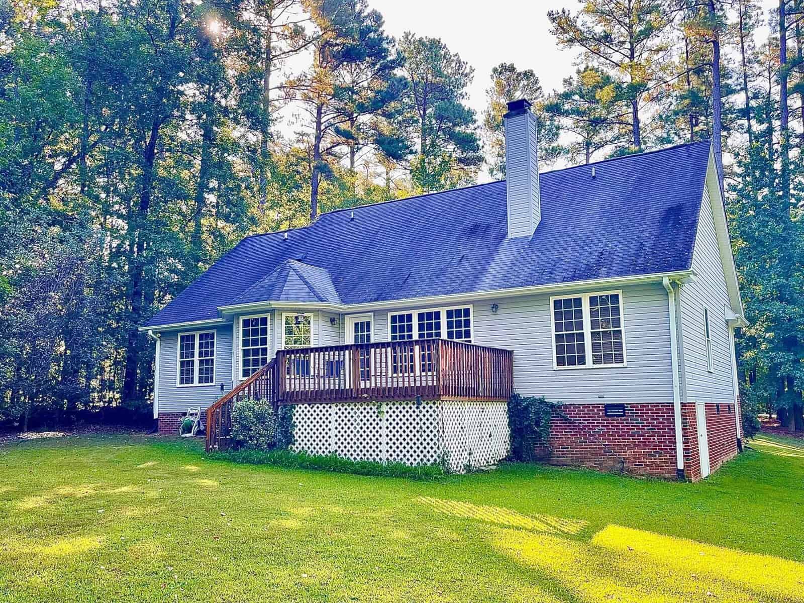 A single-story house with white siding, a dark shingled roof, and a wooden deck surrounded by tall pine trees.