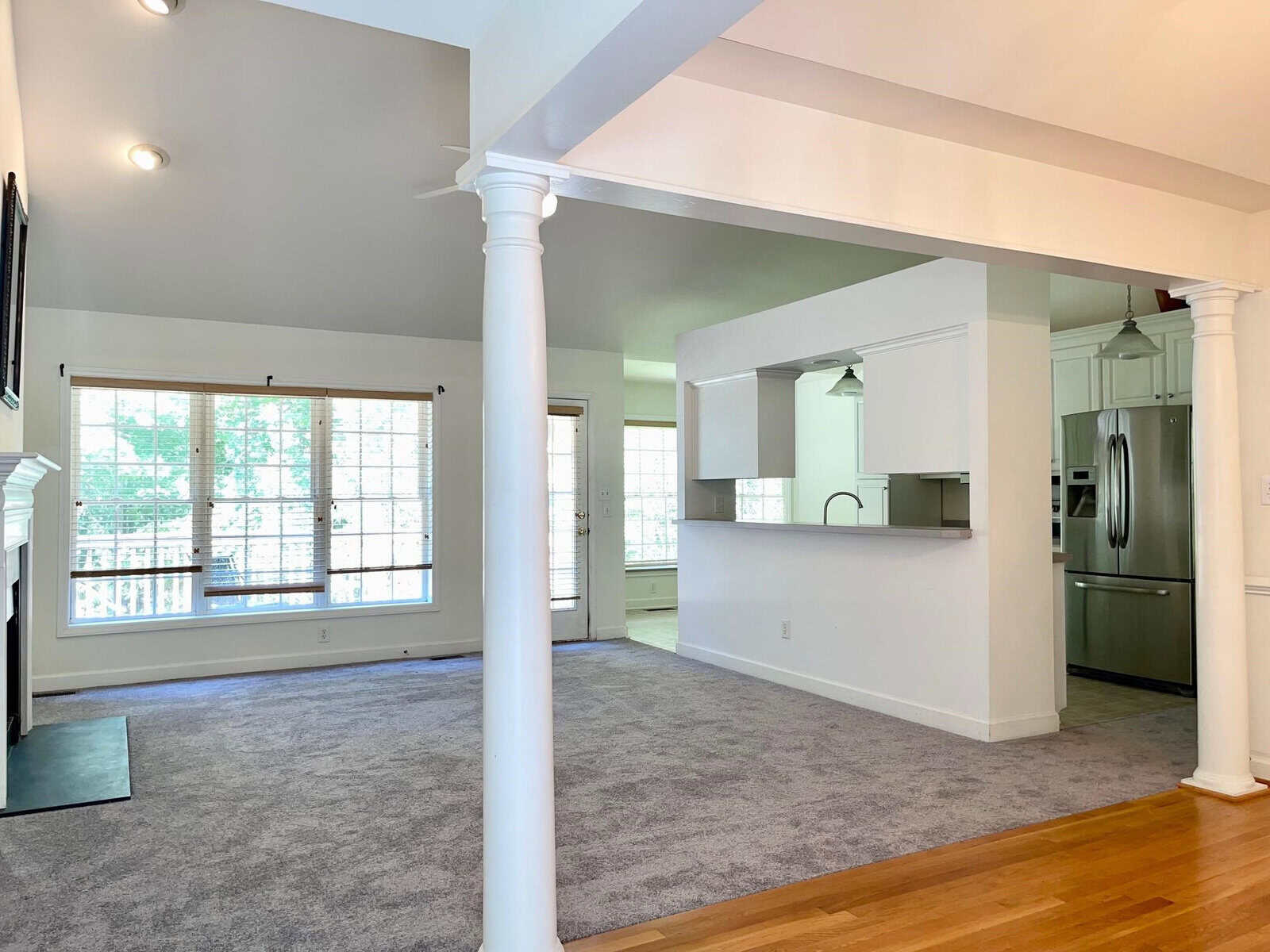 A spacious, well-lit room with gray walls and carpet flooring, featuring white columns and a view into a kitchen with modern appliances.