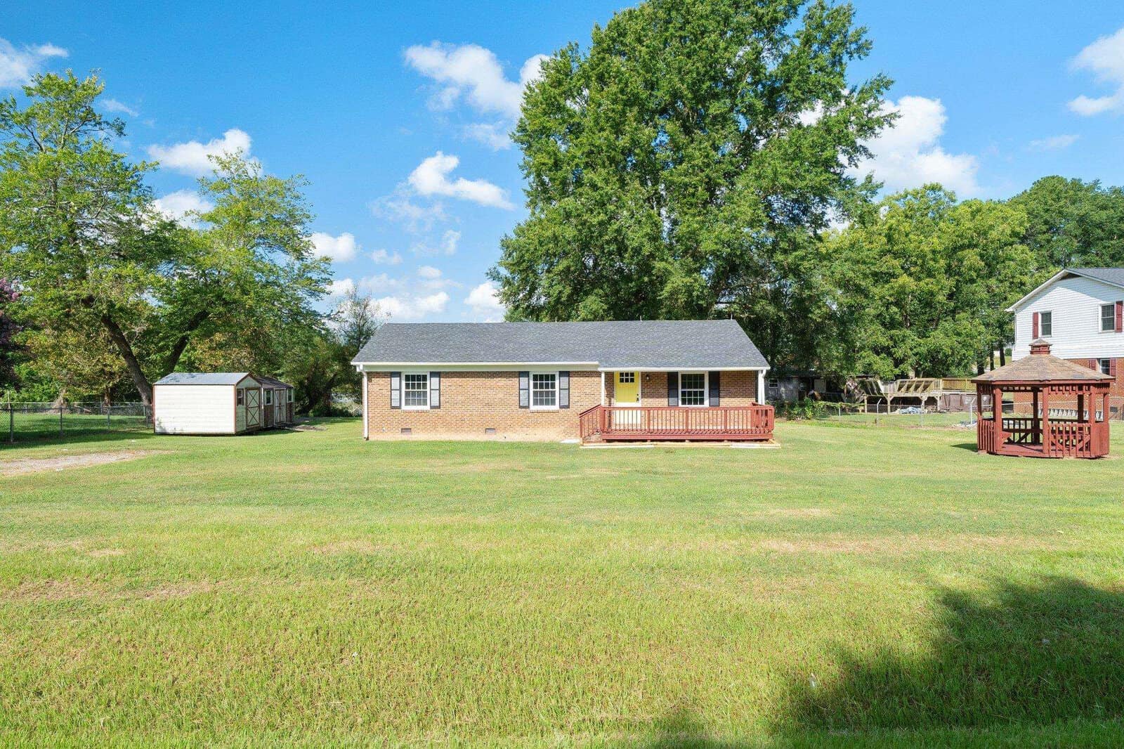 Single-story residential home with a red wooden deck and a spacious backyard featuring a gazebo and a storage shed, surrounded by a lush green lawn under a clear blue sky.
