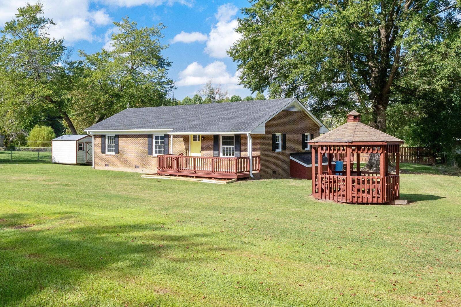A single-story brick suburban house with a red deck, surrounded by a well-kept lawn, featuring a wooden gazebo and a white storage shed in the backyard.