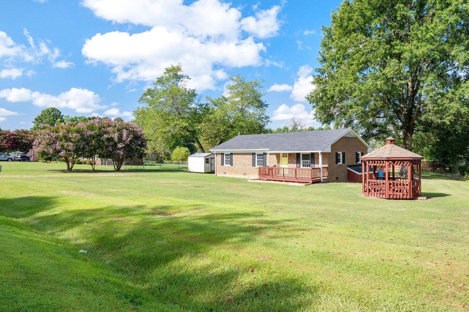 A single-story brick house with a white storage shed and a wooden gazebo, set on a large lawn with scattered trees and clear skies.
