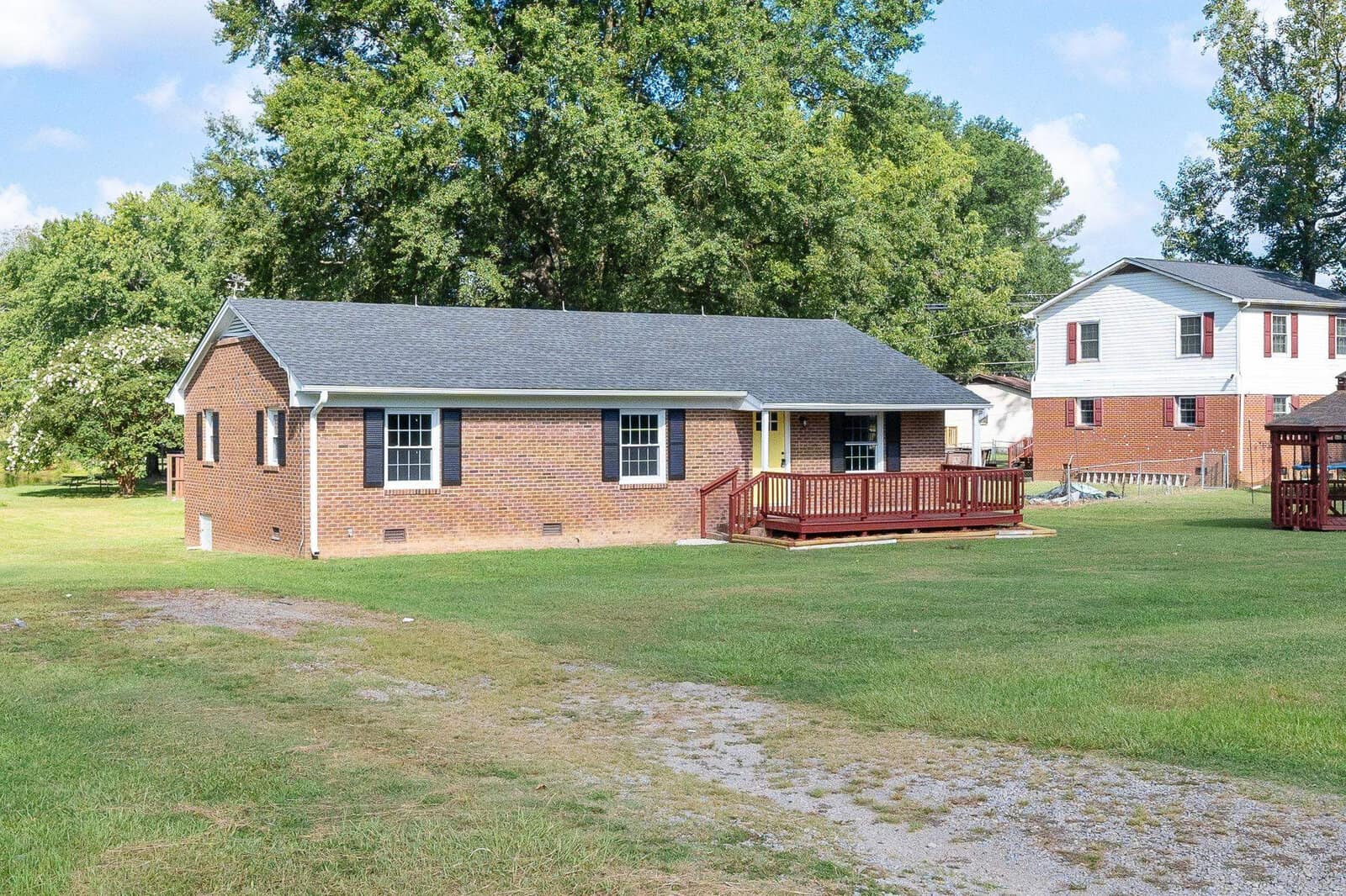 A single-story brick house with a red wooden deck and a mostly grassy front yard.