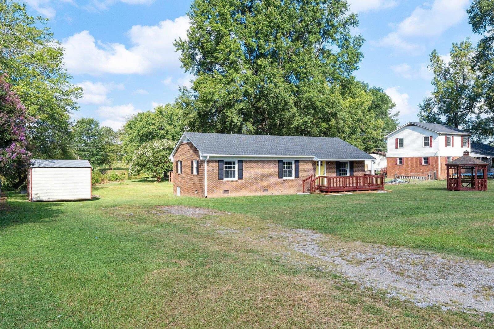 A single-story brick house with a white storage shed on the left, surrounded by a spacious lawn with a partially visible neighboring house to the right, under a partly cloudy sky.
