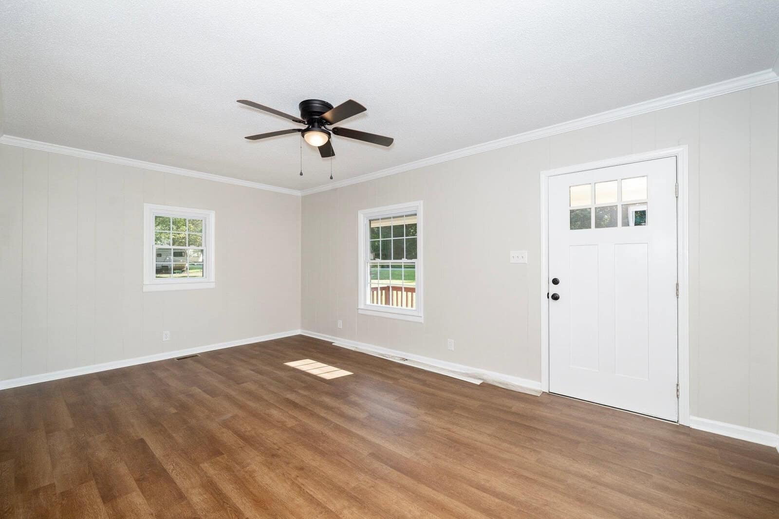An empty room with light walls, dark wooden flooring, two windows, a front door with sidelights, and a ceiling fan.