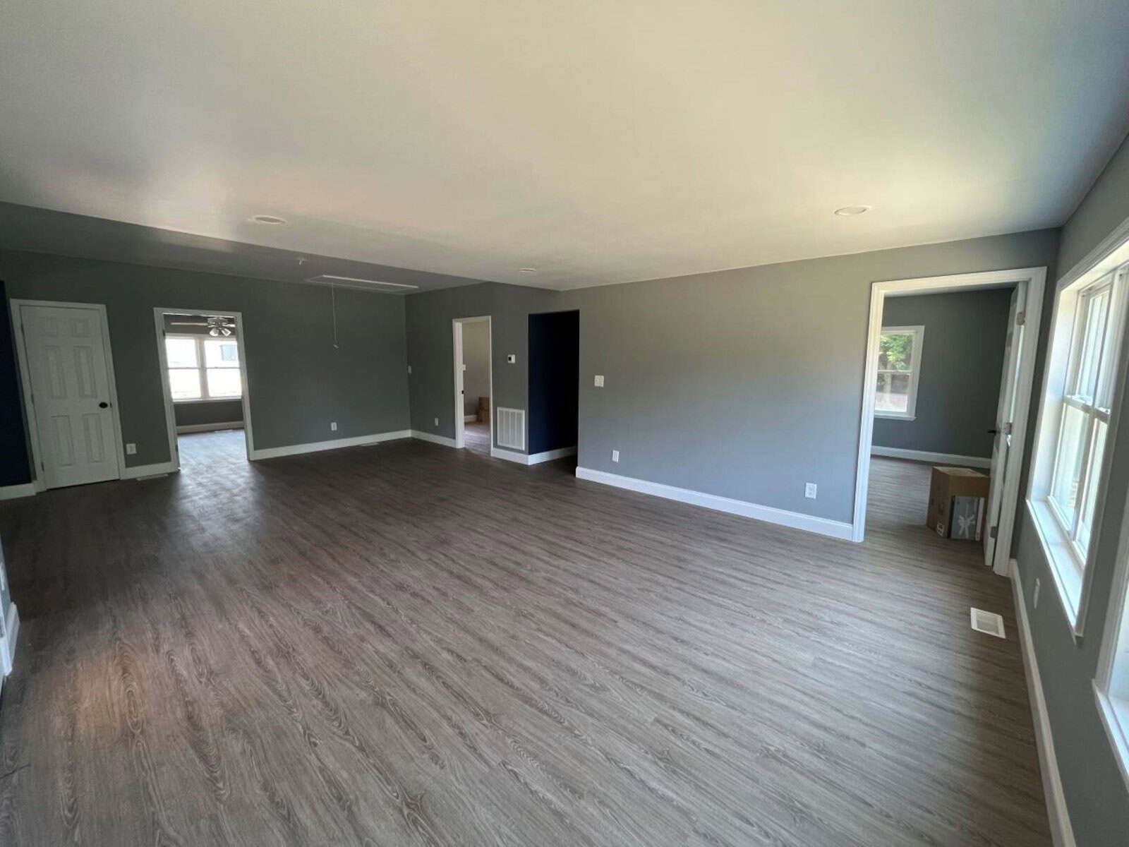 A spacious, empty room with light gray walls and wood-look flooring. Large windows let in natural light. A hallway and several doors lead to other rooms. The room has minimal furnishings, creating a clean, open feel.