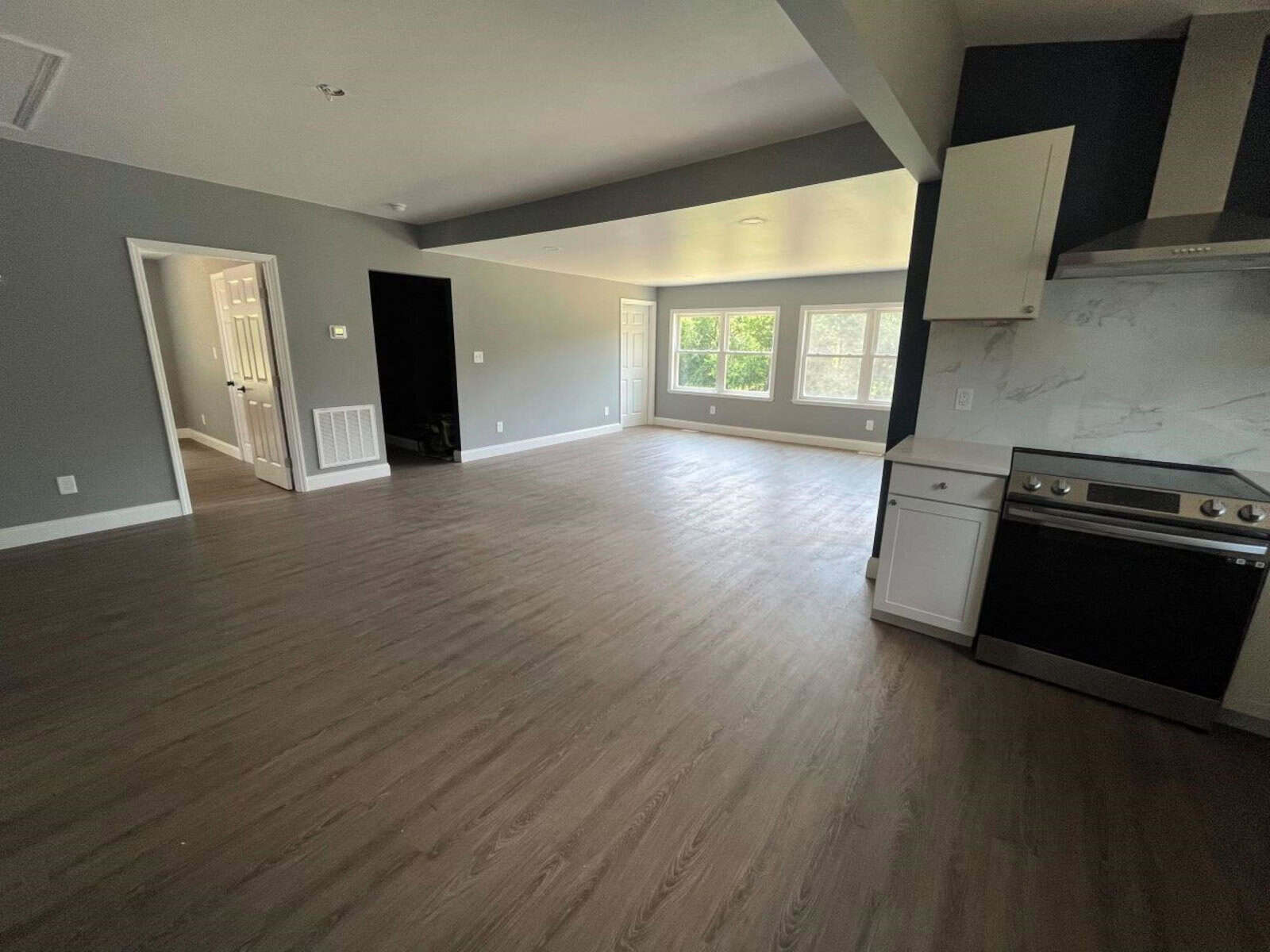 A spacious, unfurnished living area with gray walls and wood flooring. The room features large windows allowing natural light, and includes a kitchen corner with white cabinetry and a stainless steel stove. An open doorway leads to another room.